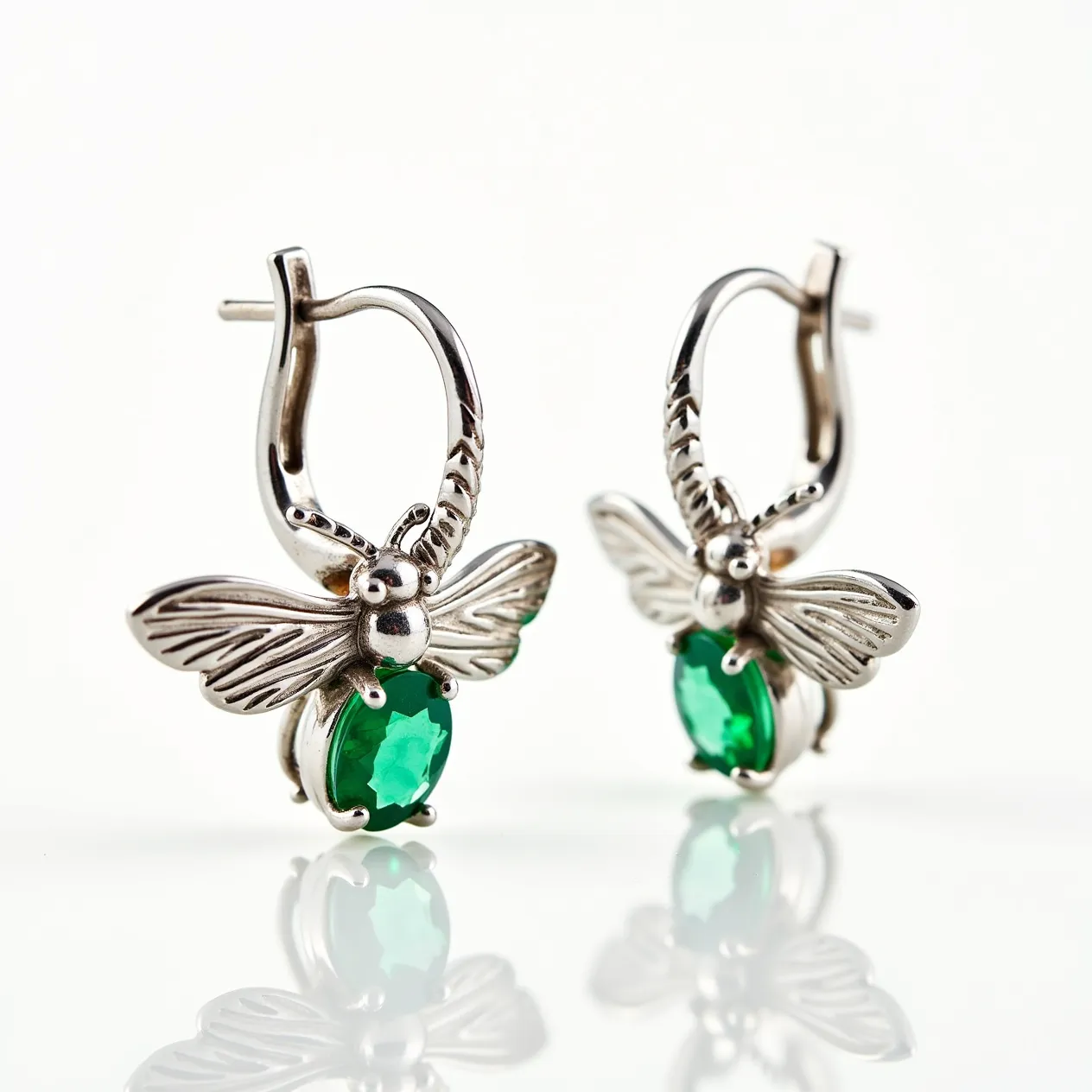 These bee earrings feature a charming design crafted from a metallic material, likely silver or a similar alloy, which gives them a sleek and polished appearance. The body of each bee is adorned with an oval-shaped green gem, possibly emerald or a synthetic equivalent, that is faceted to enhance its sparkle and set securely with prongs. The wings and antennae of the bee are intricately detailed, adding to the whimsical nature of the piece. These earrings use a latch-back clasp, providing both security and ease of wear.