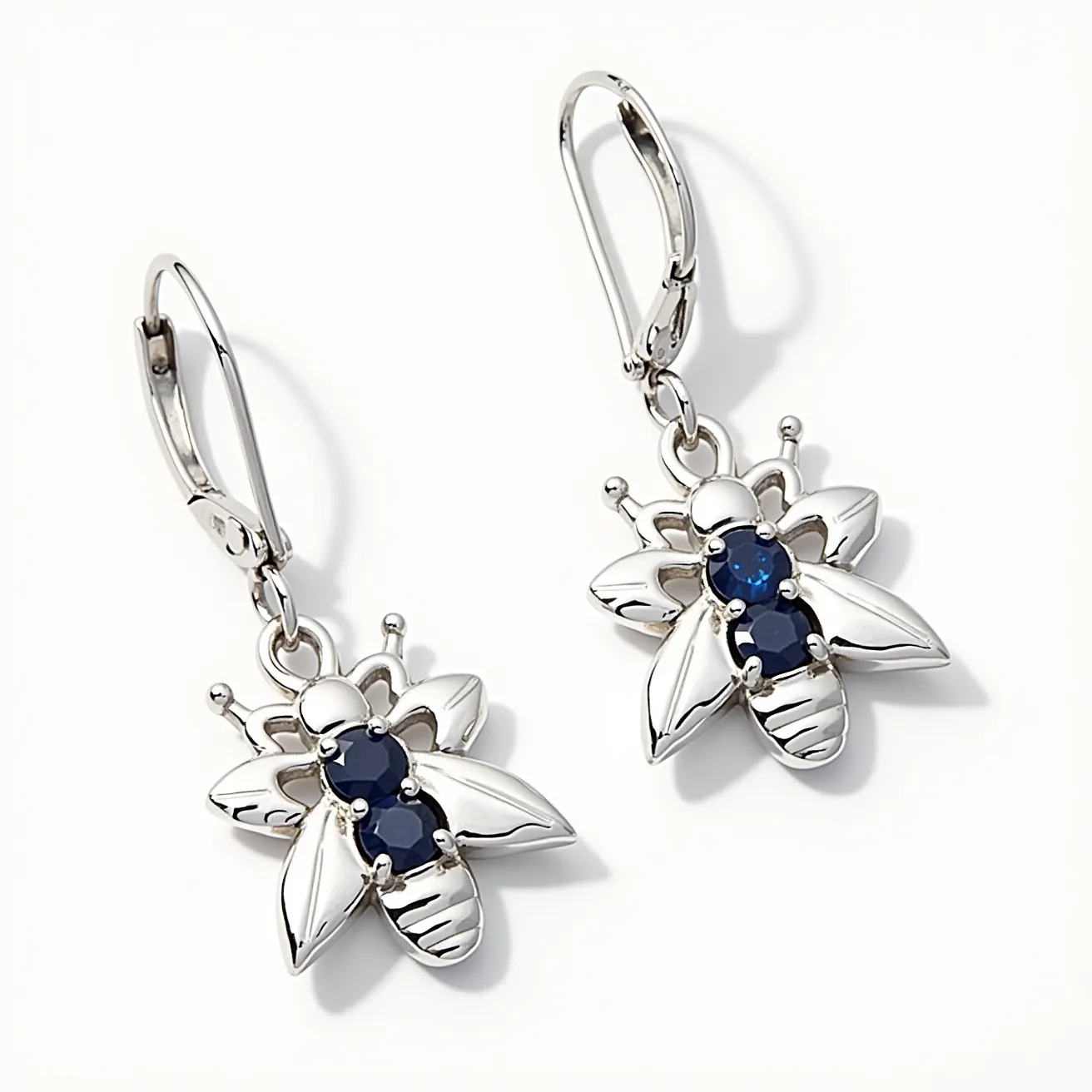 These bee earrings are crafted from a shiny metal, likely silver or a similar alloy, featuring an elegant bee design. Each bee has two round, deep blue gemstones, possibly sapphires, set in prong settings at the center of the body, adding a touch of color and sparkle. The wings and body are detailed with smooth, polished surfaces that reflect light attractively. These earrings use leverback clasps, which offer secure fastening while hanging gracefully from the ear. The design combines whimsical charm with sophisticated elegance, making them a striking accessory.