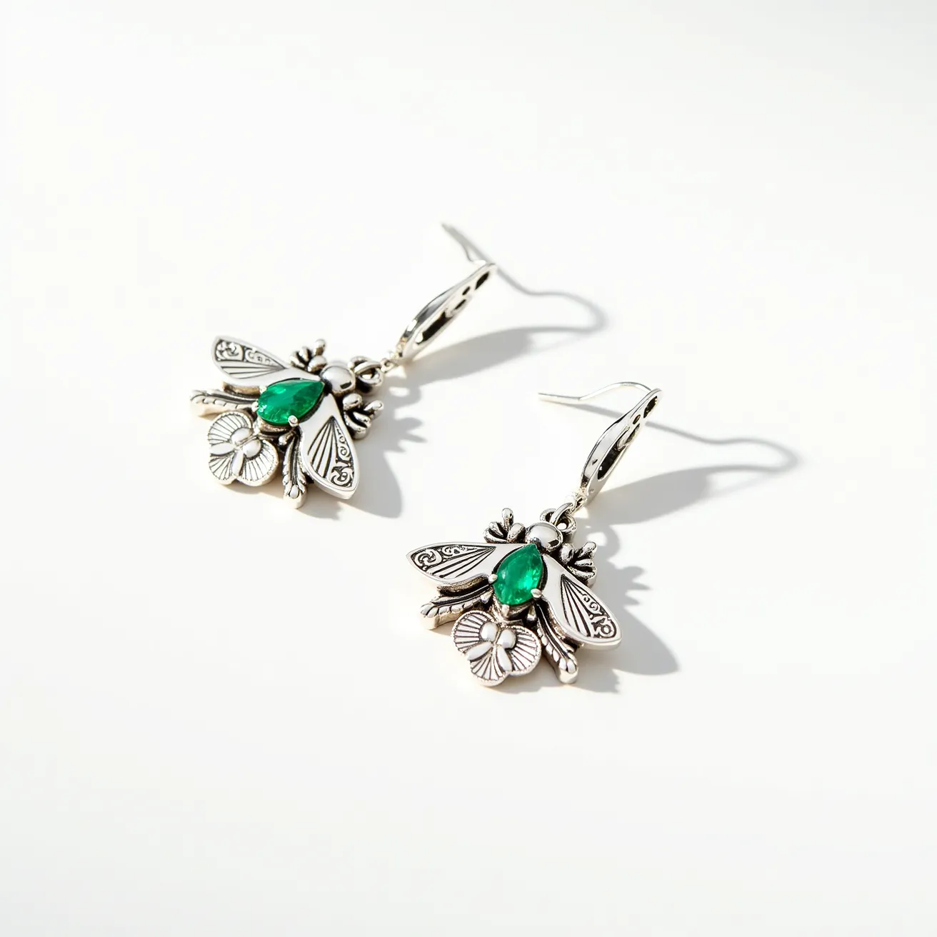 These bee earrings feature a striking design crafted from what appears to be silver metal, showcasing detailed bee motifs. Each earring is adorned with a prominent pear-shaped green gemstone, likely resembling an emerald, centrally set to represent the bee’s body. The wings and other details of the bees are intricately engraved, enhancing the overall aesthetic of the earrings. The earrings are attached via a classic hook clasp, offering ease of wear. The design combines nature-inspired motifs with elegant craftsmanship, making for a distinctive piece of jewelry.