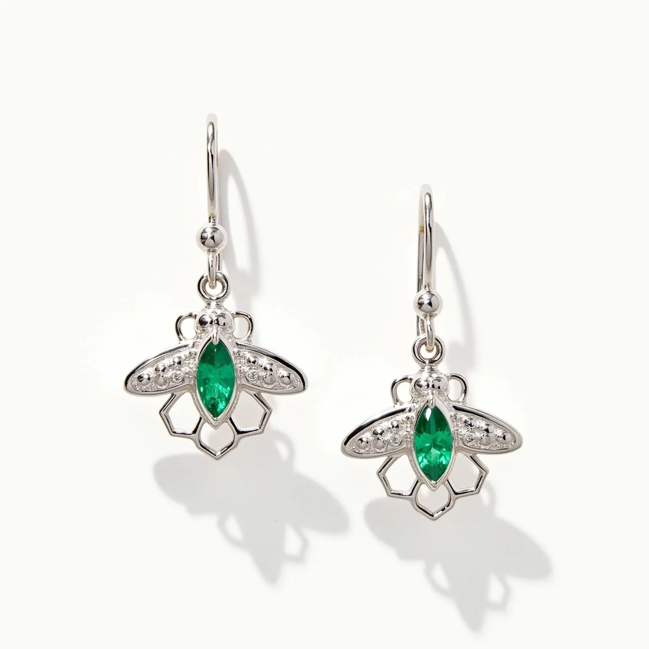 These bee earrings feature a charming design crafted primarily from what appears to be silver-toned metal. The centerpiece of each bee is adorned with a marquise-cut green gem, likely imitating the appearance of emeralds, though this is undetermined from the image alone. These gems are securely set in place, accentuating the earring's intricate design. The wings are detailed with small, round clear stones adding a touch of sparkle to the piece. The earrings are attached using hook clasps, providing ease of wear and a secure fit.