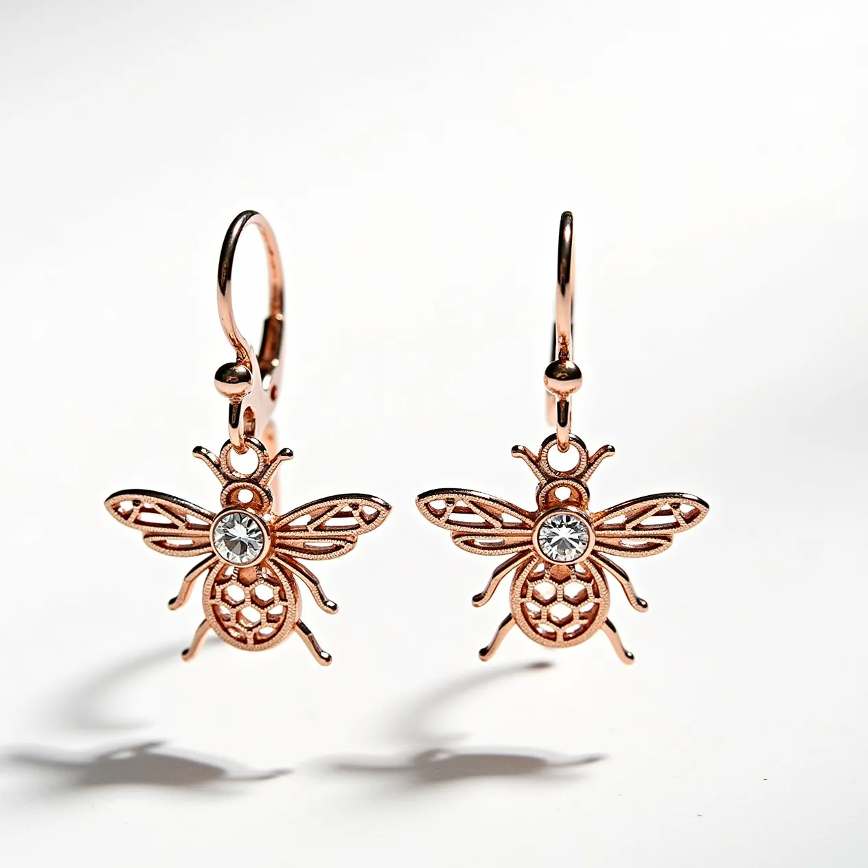 These bee earrings feature an intricate design crafted from a metal with a rose gold tone, capturing the essence of a bee with delicate, openwork detailing. At the center of each bee motif, a round, clear gemstone is positioned, possibly a rhinestone, adding a touch of sparkle and elegance. The stones are set in a bezel setting, securely holding them in place. The earrings are designed with hook clasps, providing a secure and comfortable attachment to the ear.