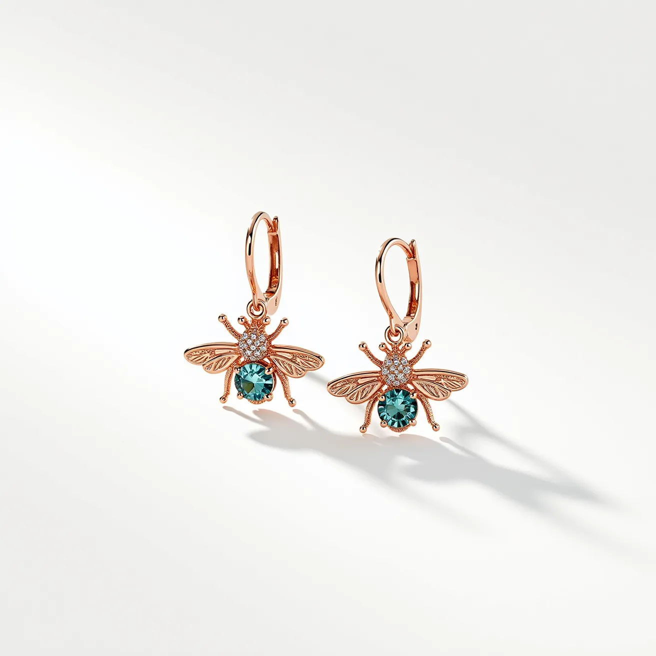 These bee earrings feature a charming design crafted from rose gold-colored metal, giving them a warm and elegant appearance. The body of each bee highlights a round, teal-colored gemstone, likely a sapphire or similarly hued stone, set in a prong setting that accentuates its brilliance. The wings and head are adorned with small, clear pavé-set stones, possibly diamonds or cubic zirconia, adding extra sparkle to the design. The earrings attach with a sleek hoop clasp, providing a secure and comfortable fit, ideal for everyday wear or a special occasion.