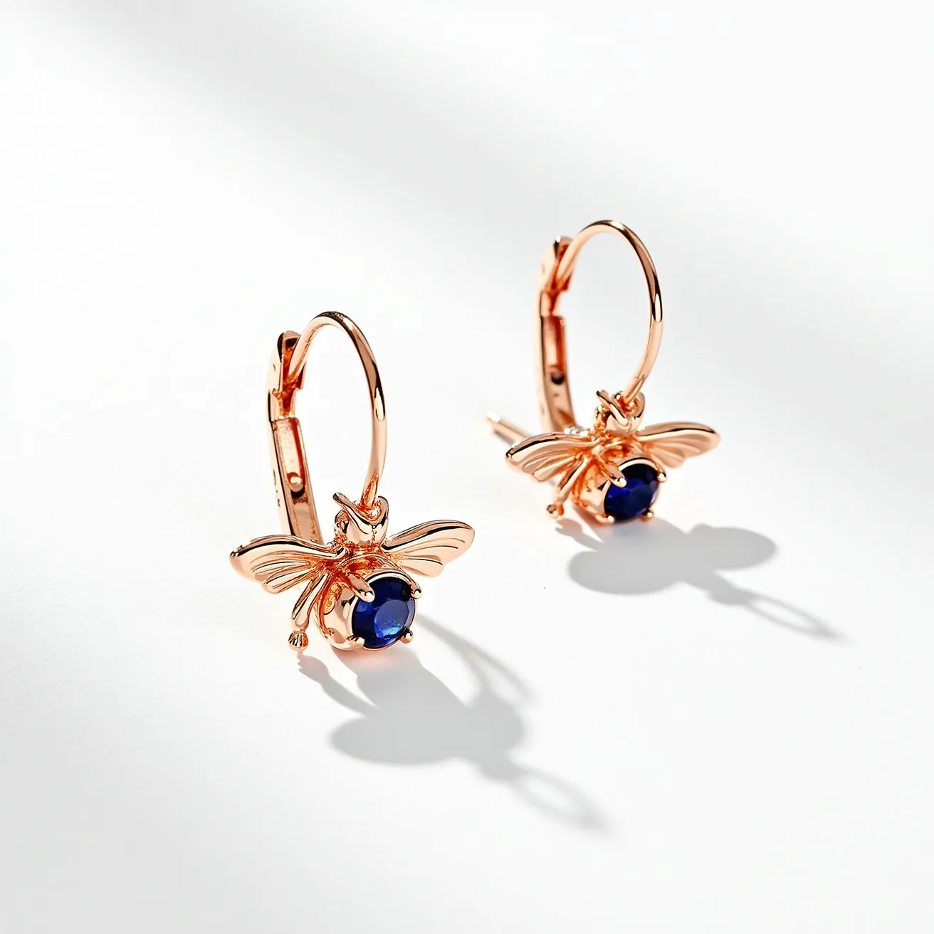 These bee earrings feature an elegant design crafted from a warm-toned metal, likely rose gold, creating a delicate bee shape. Each earring showcases a single deep blue gemstone, possibly a sapphire, which is round cut and securely held in place by a prong setting, adding a touch of sophistication and contrast to the design. The earrings are attached to large hoops, offering a secure and classic lever-back clasp that ensures ease of wear and a comfortable fit. The combination of the vibrant gemstone and detailed metalwork highlights the fine craftsmanship and adds a touch of nature-inspired elegance to the piece.