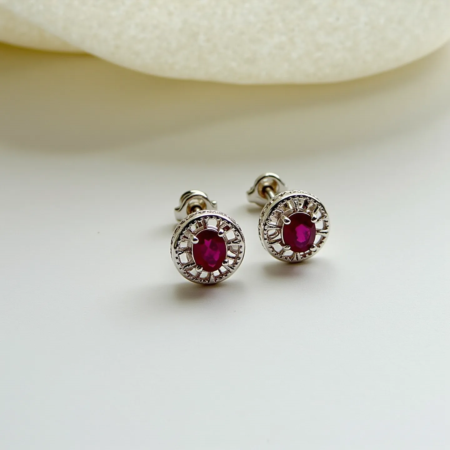 These birthstone earrings feature a captivating design centered around vivid red gemstones, likely rubies, which are intricately cut and set in a classic prong setting. The stones are surrounded by a delicate halo of smaller clear gems, enhancing their vibrant hue and adding a touch of elegance to the overall design. Crafted from a silvery metal, possibly sterling silver or white gold, the earrings exhibit a refined and polished finish. The attachment is a secure post-back style, ensuring a comfortable and stable fit for the wearer.