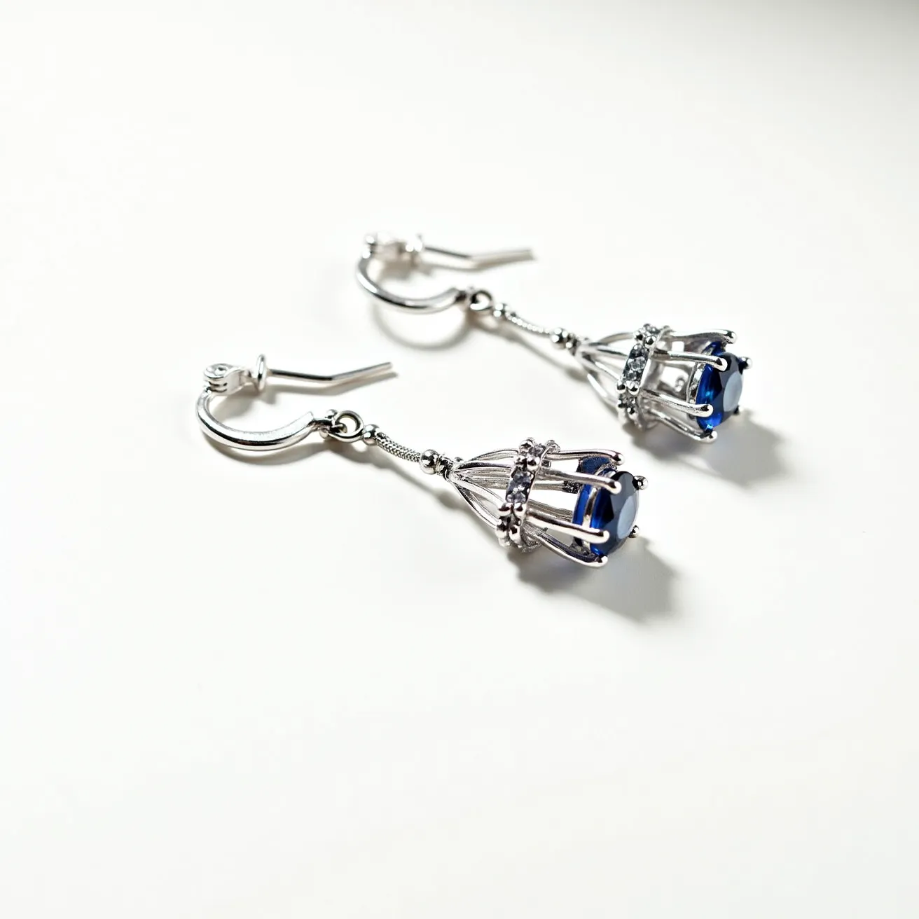 These birthstone earrings feature a striking blue gemstone, possibly representing sapphire, which is often associated with the month of September. The gem is skillfully cut to enhance its vibrant color and is securely held in a unique cage-like setting made of what appears to be a silver-toned metal. The setting includes small decorative elements that add an extra touch of elegance to the design. The earrings are equipped with hook clasps that allow for easy and secure attachment, making them both stylish and practical.