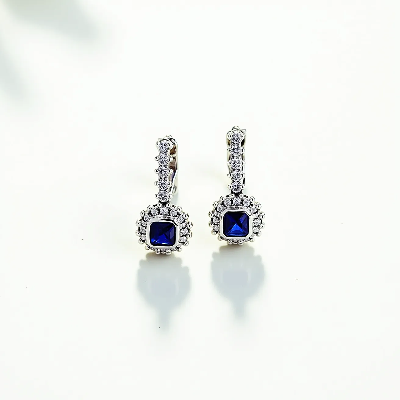 These birthstone earrings feature a central square-cut deep blue gemstone, likely sapphire, surrounded by a halo of smaller clear stones, possibly diamonds, in a pavé setting. The earrings appear to be crafted from a shiny material, likely white gold or platinum, enhancing their elegant appearance. The attachment is a leverback clasp, ensuring a secure fit while complementing the decorative design of the piece. The overall style is sophisticated and classic, making these earrings suitable for various occasions.