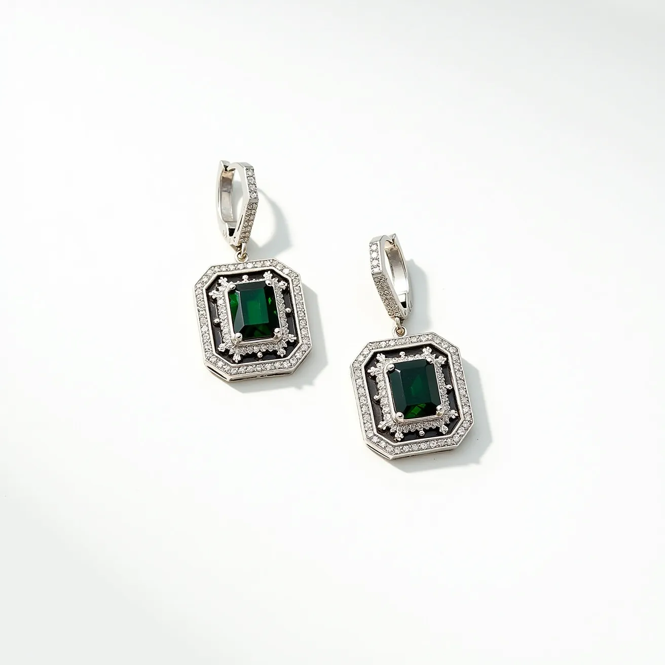 These black earrings feature a striking design with an emerald-cut dark green gemstone centrally located in each piece, which is encased in a symmetrical, geometric setting. The stones are bordered by two rings of black material, possibly onyx or enamel, adding depth and contrast to the piece. Surrounding the black accentuations is a row of small, clear gemstones, likely diamonds, arranged in a pavé setting for added brilliance. The earrings attach with a lever-back clasp, ensuring a secure and comfortable fit. The use of a polished metal, possibly white gold or platinum, offers a refined finish, complementing the overall elegant aesthetic.