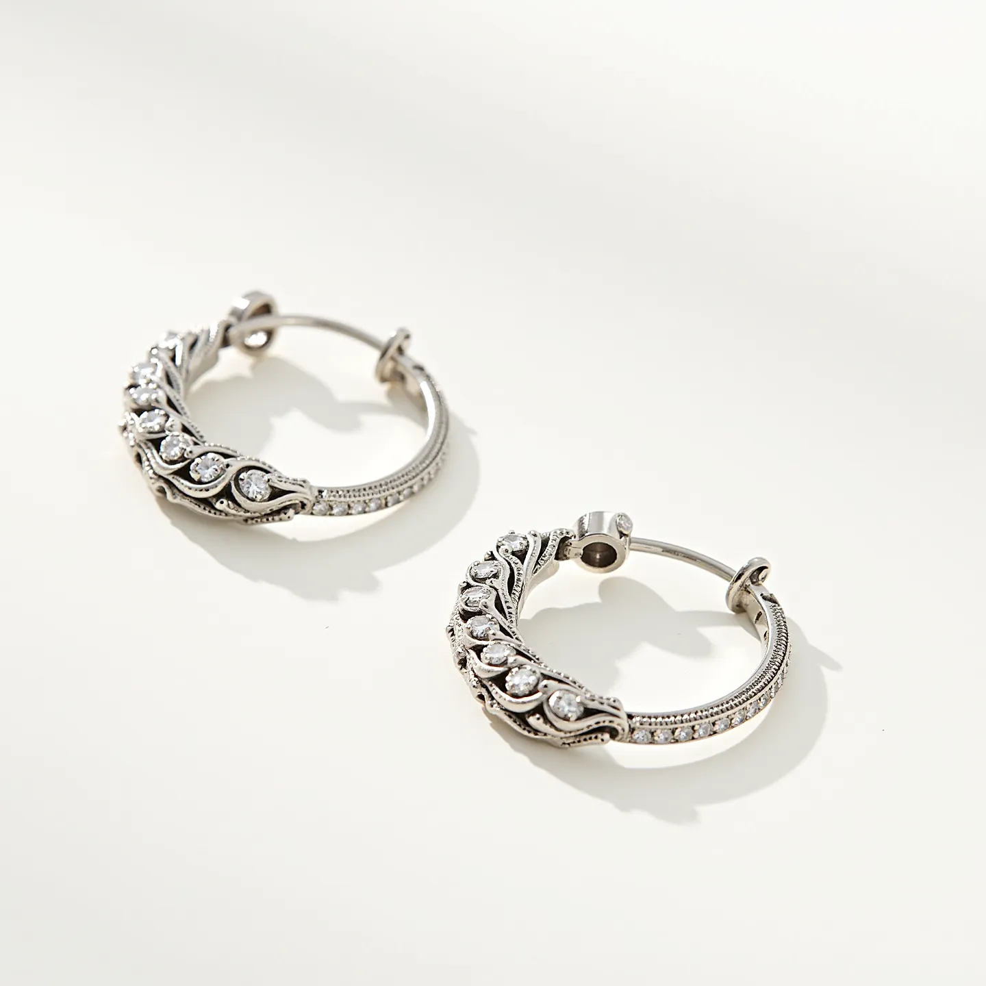 These cartilage hoop earrings are crafted from a shiny metal, likely silver or white gold, featuring an intricate design with decorative elements. The hoops are adorned with a sequence of round-cut clear stones, possibly cubic zirconia or diamonds, set securely within a detailed metal border that adds a sense of elegance to the piece. The earrings include a hinge mechanism with a secure clasp, ensuring easy attachment and a comfortable fit on the earlobe, fitting snugly to hug the cartilage area stylishly.