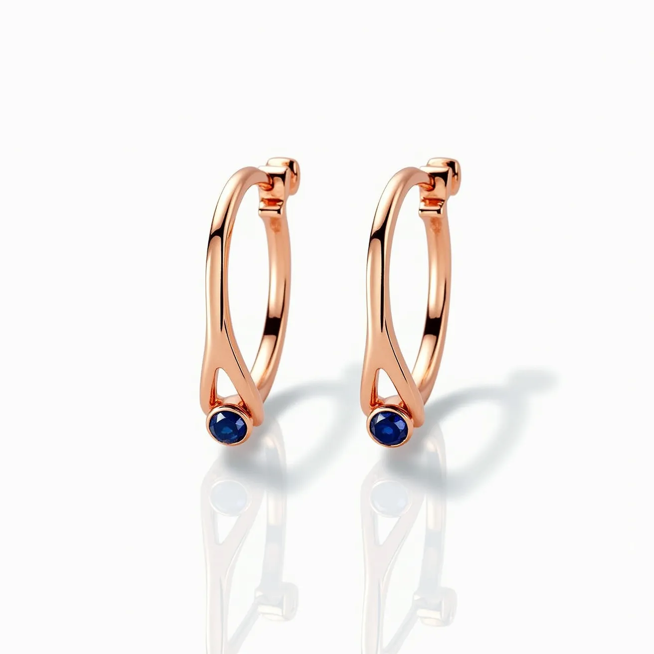 These cartilage hoop earrings are crafted from a rose gold metal, offering a sleek and modern look. Each hoop features a small, round blue gemstone set at the base, likely a sapphire, adding a touch of elegance. The stone is set in a bezel setting, which securely encases and enhances the gem's natural beauty. The hoops utilize a hinge clasp mechanism, ensuring ease of use and a secure fit.