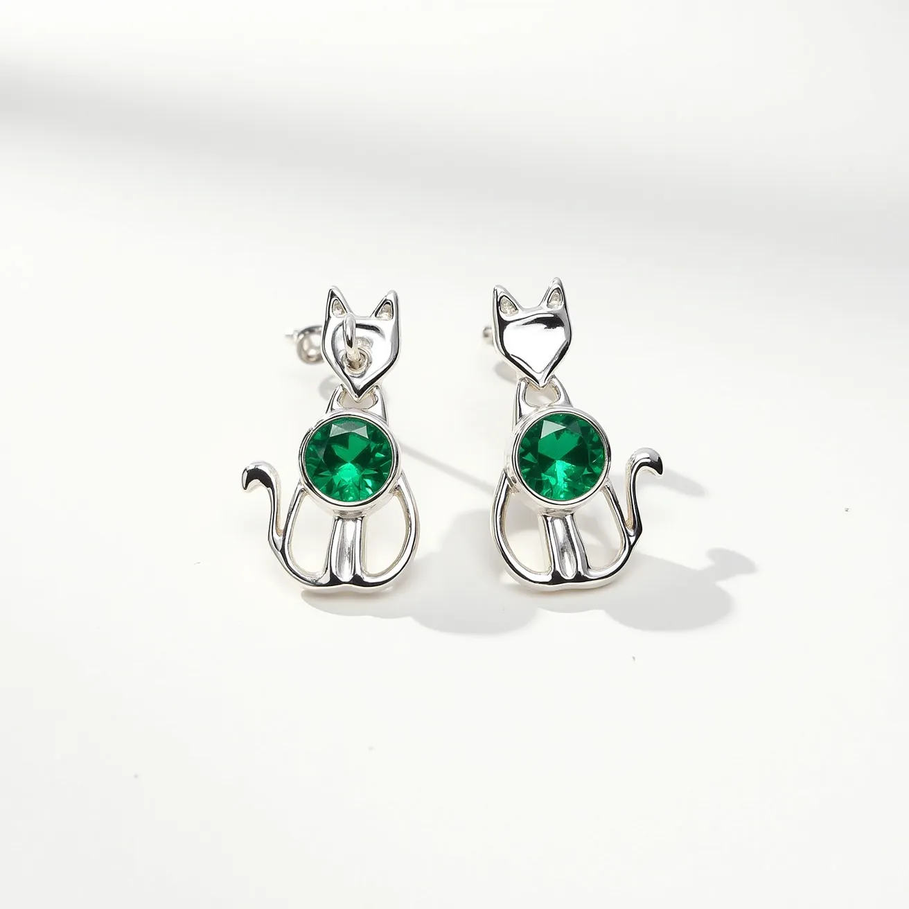 These cat earrings feature a sleek, silver-toned metal design shaped into a minimalist silhouette of a cat. At the center of each earring is a striking green gem, cut with facets to catch the light, and securely set in a bezel setting that complements the metallic structure. The earrings are designed with a post and push-back clasp, ensuring a secure attachment when worn.