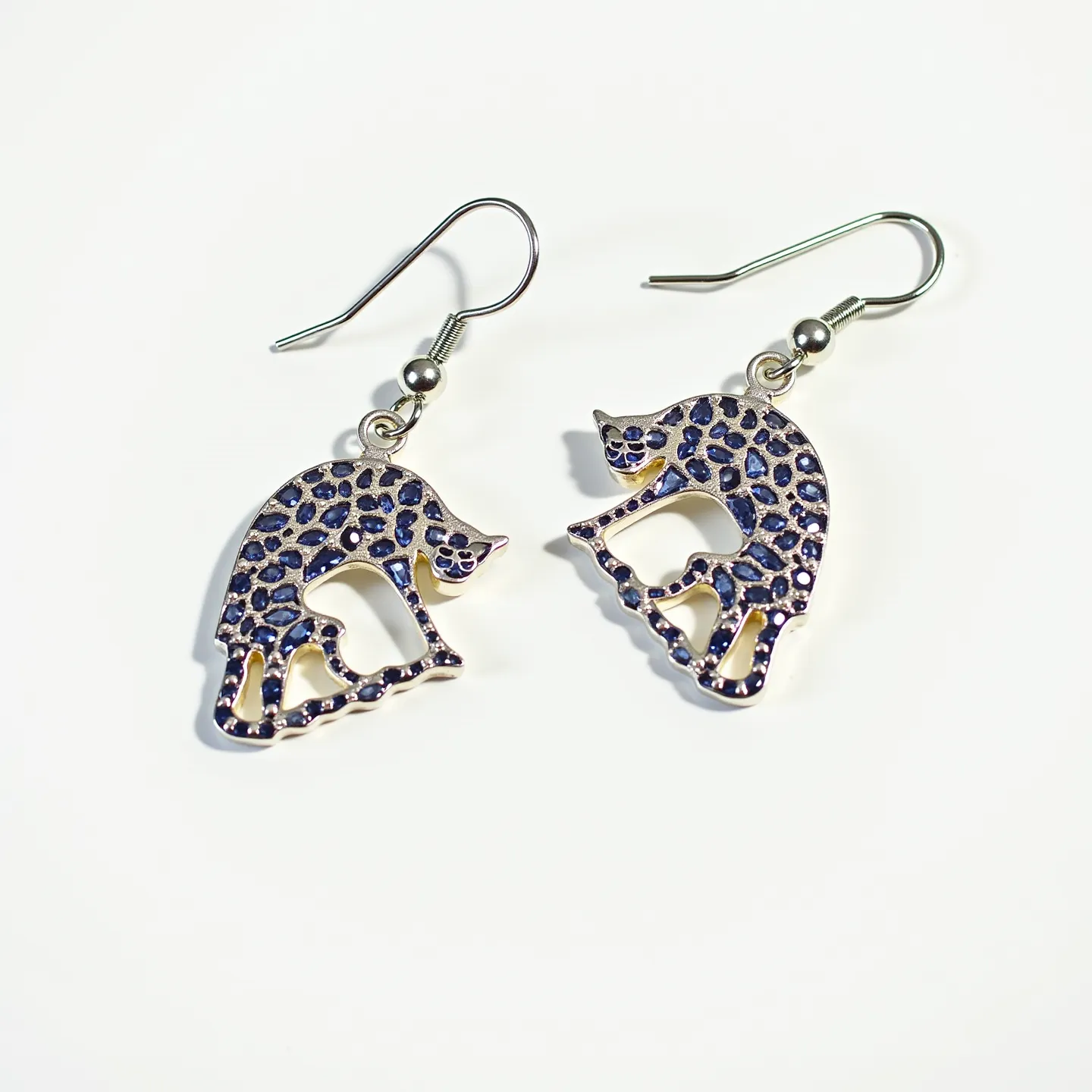 These cat earrings feature a charming design with cats intricately crafted from a metal that appears silver in tone. The cat figure is adorned with a pattern resembling dark blue stones, giving an impression of a spotted texture. These are likely imitation stones, set flush within the surface, contributing to a playful and stylish look. The earrings are attached to a simple hook clasp, allowing for easy and secure wear. The overall design combines whimsy with elegance, ideal for casual or themed occasions.