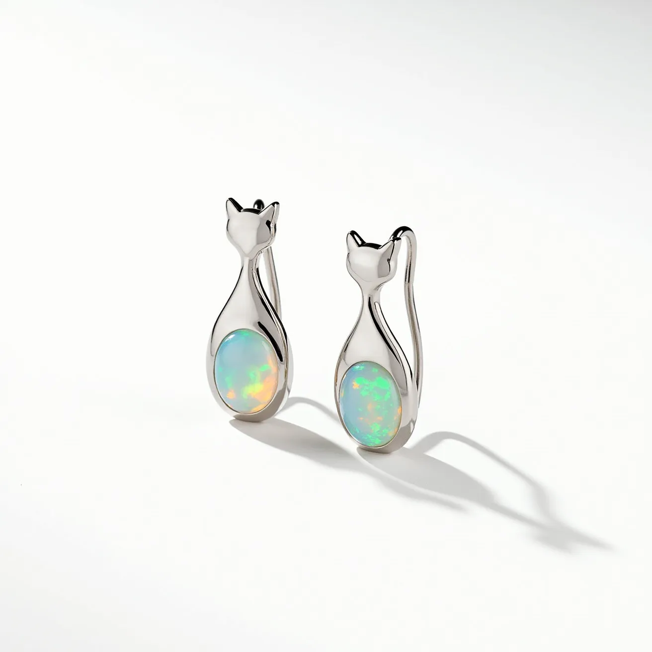 These cat earrings feature a sleek, polished metal design shaped like a stylized cat silhouette. At the body of each cat is an oval opal gemstone, displaying vibrant, multicolored hues that catch the light. The opal appears to be bezel-set, providing a smooth and continuous frame around the stone that integrates seamlessly with the overall design. These earrings likely use a simple hook attachment, complementing their elegant and minimalist appearance. The combination of the opal's iridescence and the shiny metal creates a striking visual appeal.