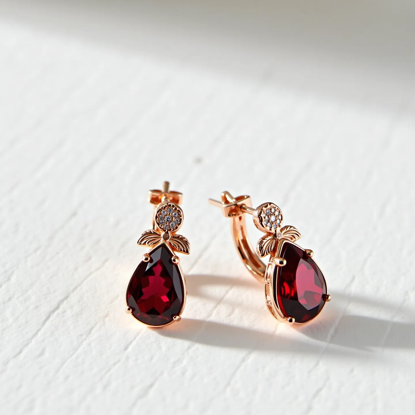 These cherry earrings feature a striking design with large, teardrop-shaped red stones, likely garnets or similar, set in a gold-tone metal. The faceted stones are securely held in a classic prong setting. Above each stone, there is a decorative element resembling leaves crafted from the same gold-tone metal. Small round white gems, possibly diamonds or cubic zirconias, embellish the top part of the earrings, adding sparkle. The earrings are equipped with latch back clasps, ensuring a secure fit when worn.