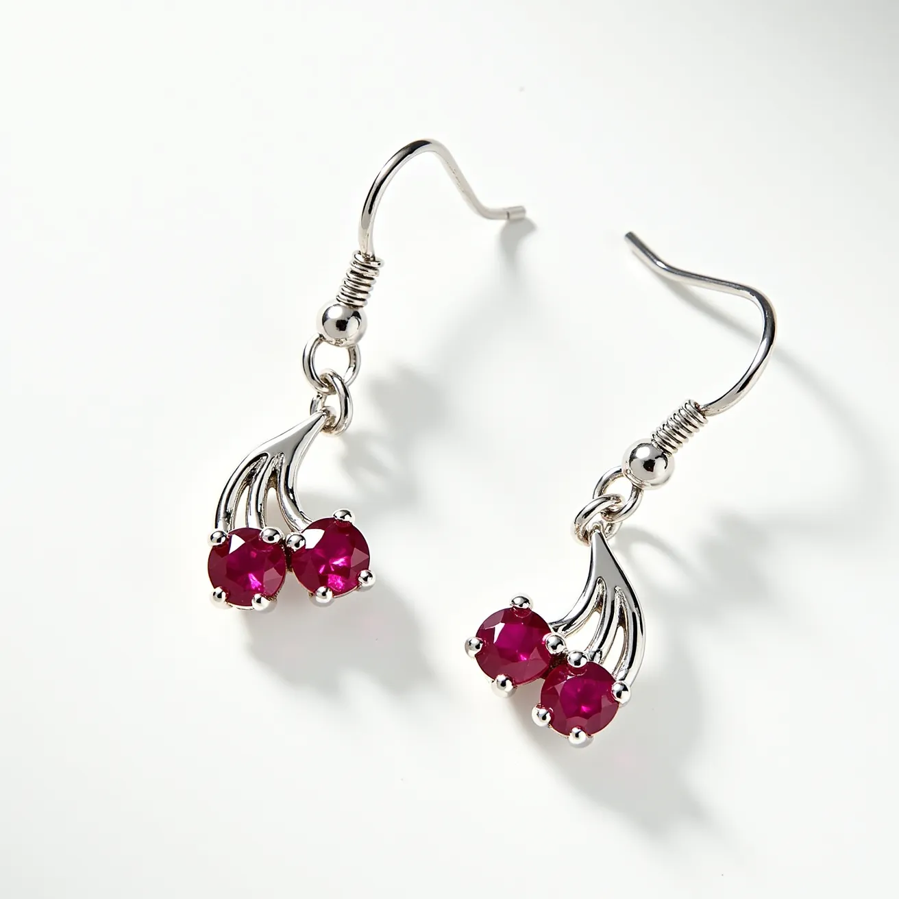 These cherry earrings feature a delicate design crafted from a silver-toned metal, forming a stylized cherry motif. Each earring showcases two round-cut red gemstones that resemble cherries, securely set in four-prong settings. The stones are arranged in pairs, attached to the end of elegant, curved stems that enhance the cherry appearance. These earrings are designed with hook clasps, adding both elegance and ease of wear. The combination of the red gemstones and silver-toned metal creates a striking and vibrant aesthetic, making them a charming accessory.