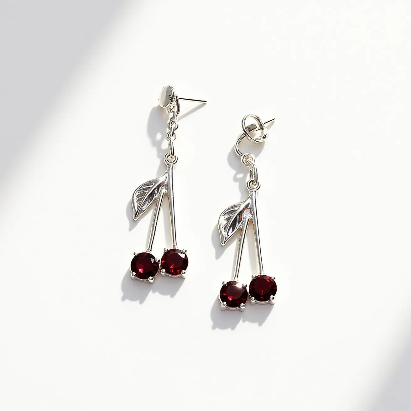 These cherry earrings feature a playful and elegant design with two deep red gemstones resembling cherries as the focal point. The stones appear to be round-cut and are set securely in a prong setting that enhances their brilliance. Each "cherry" is connected to thin, graceful metal stems, crafted from what looks like silver, leading up to a leaf detail, also in silver, which adds a touch of nature to the design. The earrings are equipped with a post backing, providing a secure and comfortable fit for pierced ears, while maintaining a sleek and stylish aesthetic.