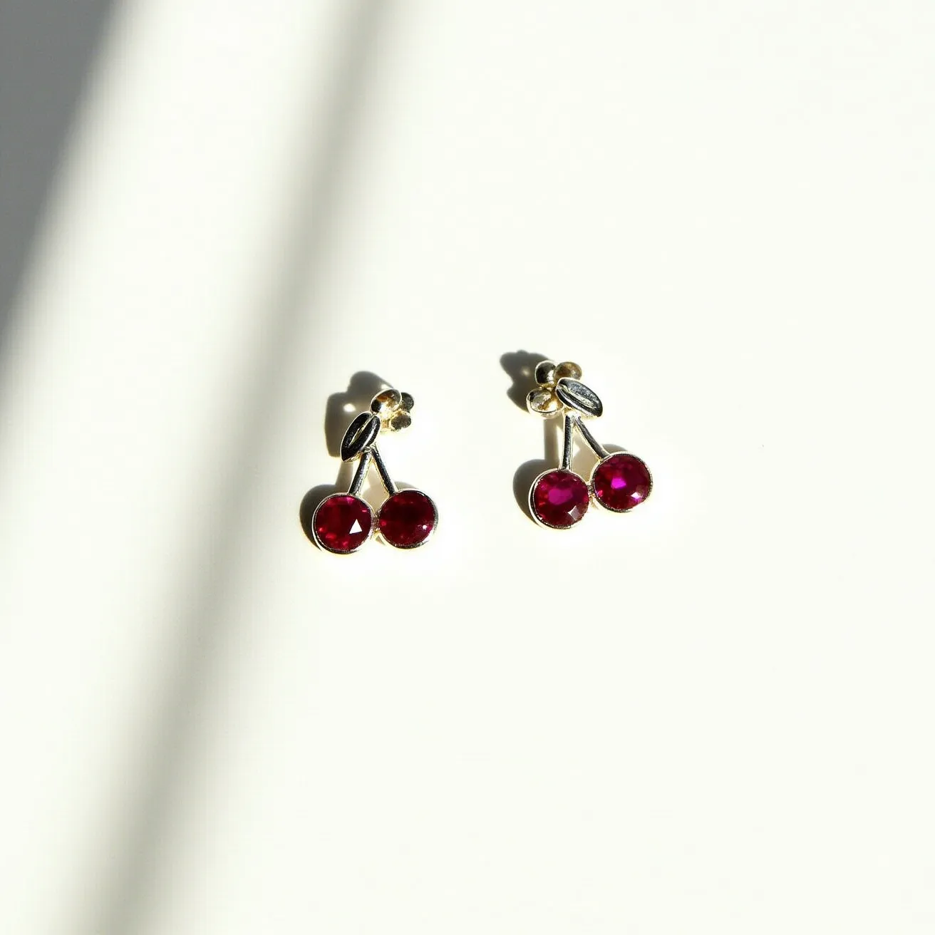 These cherry earrings feature two bright red, round-cut stones set in each earring, resembling cherries. The stones are likely glass or synthetic gems and are set in a gold-toned metal. Each pair of cherries is connected to a small gold-toned stem with a single leaf detail, adding a realistic touch. The earrings appear to have a stud backing, providing a secure and comfortable fit for pierced ears.