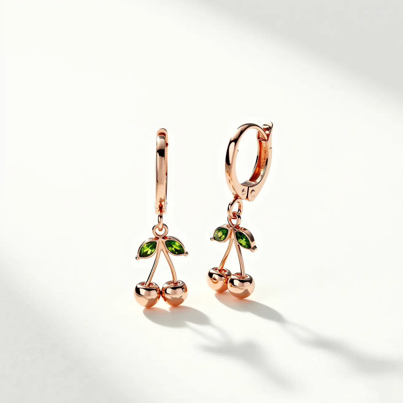 These cherry earrings feature a playful design crafted from a rose gold-toned metal, highlighting a charming cherry motif. The piece includes a pair of lush green gems, cut in a marquise shape, set delicately as leaves above the cherries, adding a vibrant sparkle to the earrings. The cherries themselves are formed by polished metal spheres, achieving a sleek and glossy look. The earrings are secured with hinged hoop clasps, ensuring ease of wear and a secure fit, while maintaining the elegant and whimsical aesthetic.