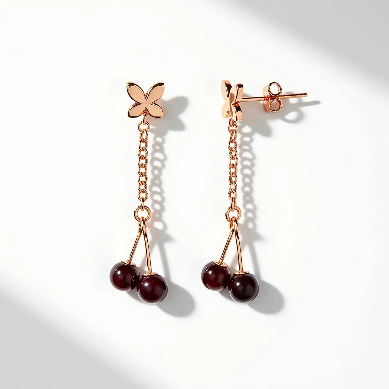 These cherry earrings feature a charming design with two deep red, spherical beads that resemble cherries. They are suspended delicately from a sleek and elegant gold setting. The beads are likely made from a polished stone or synthetic material, accentuating a glossy and vibrant appearance. The gold chain attachment adds a refined touch and connects to a flower-shaped stud, adding a playful yet sophisticated element to the design. The earrings are secured with a classic post and butterfly clutch back that ensures a comfortable and secure fit.