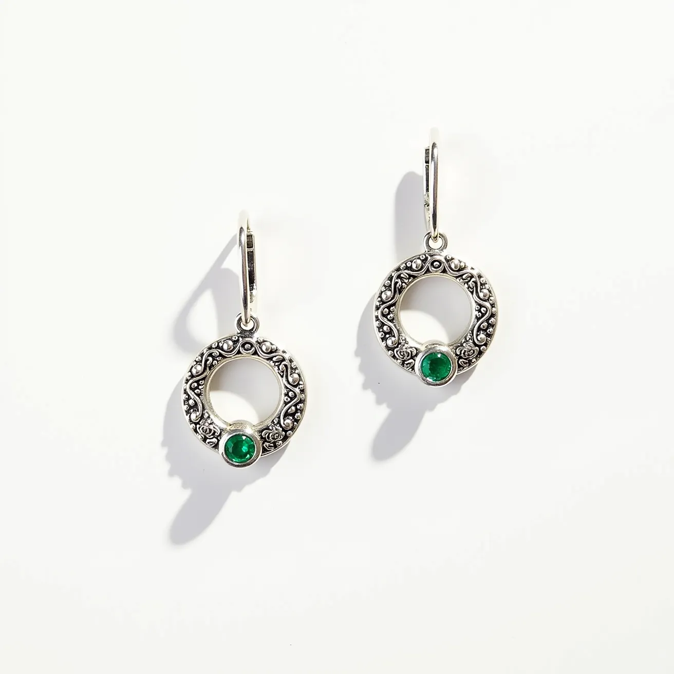 These circle earrings feature an intricate design made of silver, showcasing ornate detailing around the circular frame. At the base of each earring, a round, green gemstone is set, likely an emerald, which is bezel set to ensure durability and elegance. The earrings dangle from lever-back clasps, providing a secure and comfortable attachment for the wearer. The combination of the vivid green stones with the detailed silver work creates a classic and elegant look, perfect for adding a touch of sophistication to any outfit.