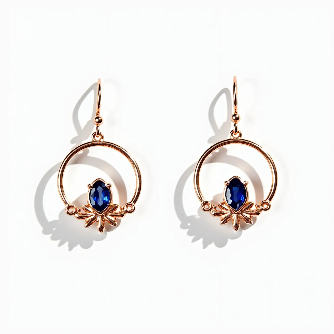 These circle earrings feature a delicate gold-toned metal hoop design, accented by a central marquise-cut blue gemstone, possibly a sapphire, set prominently within a prong setting. The gemstone is flanked by intricate leaf-like motifs, adding an elegant touch to the overall appearance. These earrings are designed with a hook clasp that allows for easy attachment and graceful dangle, contributing to their sophisticated look. The luxurious combination of the rich blue stone and the warm gold setting creates a striking and timeless aesthetic.