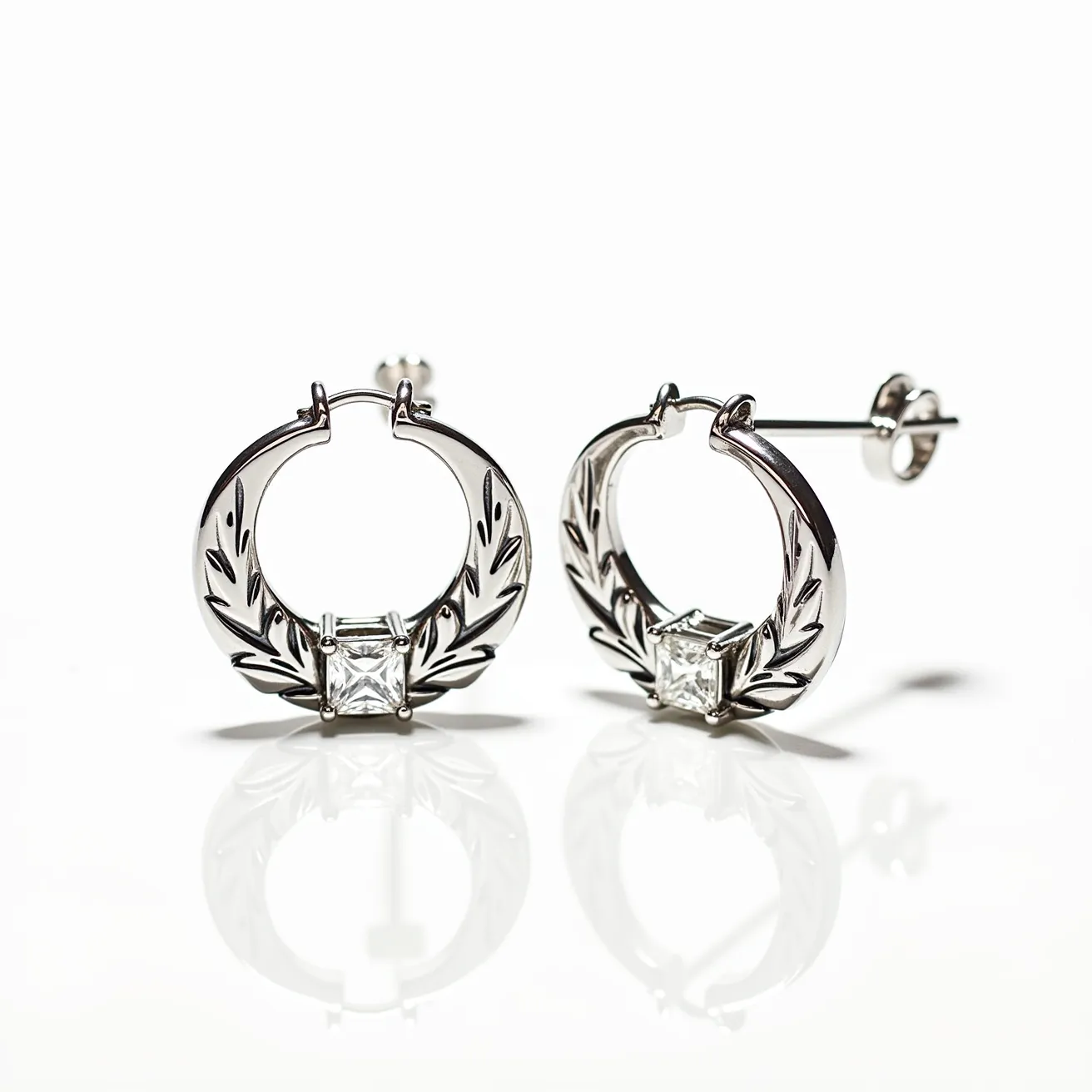 These circle earrings are crafted from a lustrous silver-toned metal, featuring an intricate leaf-like design engraved onto the hoops. Each earring showcases a prominent square-cut gem, likely a clear stone, securely held in place by a four-prong setting at the base. The earrings have a post and butterfly clasp, ensuring a secure fit when worn. The combination of the shiny metal and the central stone creates an elegant and refined appearance.