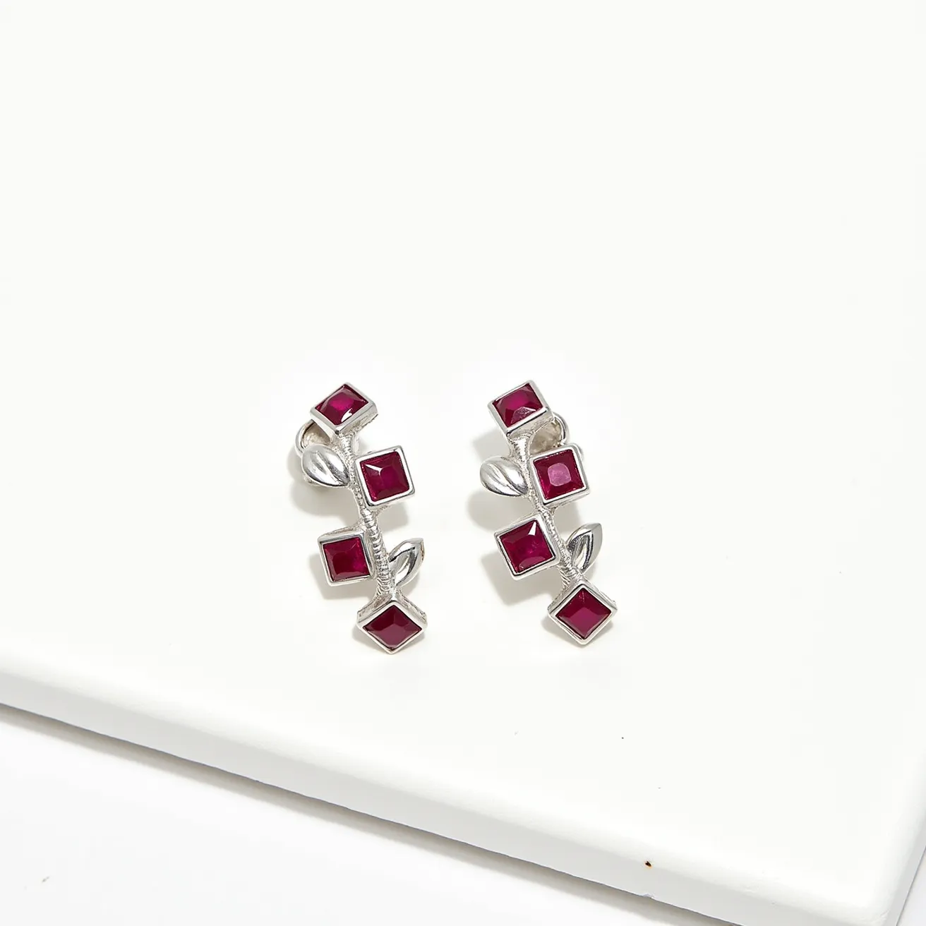 These clip on earrings feature a chic and elegant design, crafted primarily from a polished silver-toned metal. They are adorned with square-shaped, deep red stones, likely rubies or ruby-like gems, each set in a sleek bezel setting that enhances their vibrant color. The earrings have a leafy motif with small, silver decorative leaves interspersed between the stones, adding a touch of nature-inspired grace. The clip-on mechanism enables easy attachment without the need for pierced ears, making them a versatile choice for various occasions.