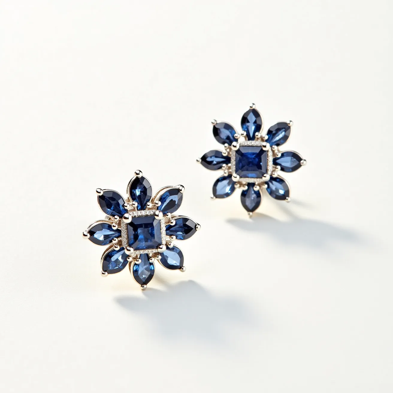 These clip on earrings feature a distinctive floral design, crafted with a central square-cut sapphire-like gemstone secured within a prong setting. Surrounding the central stone, eight marquise-cut stones radiate outward, also of a sapphire-like hue, enhancing the overall aesthetic with their rich blue tones. The stones are set in a polished metal, likely silver or white gold, which provides a striking contrast to the deep blue of the gems. The attachment mechanism for the clip-on earrings is not visible, ensuring a seamless appearance when worn.