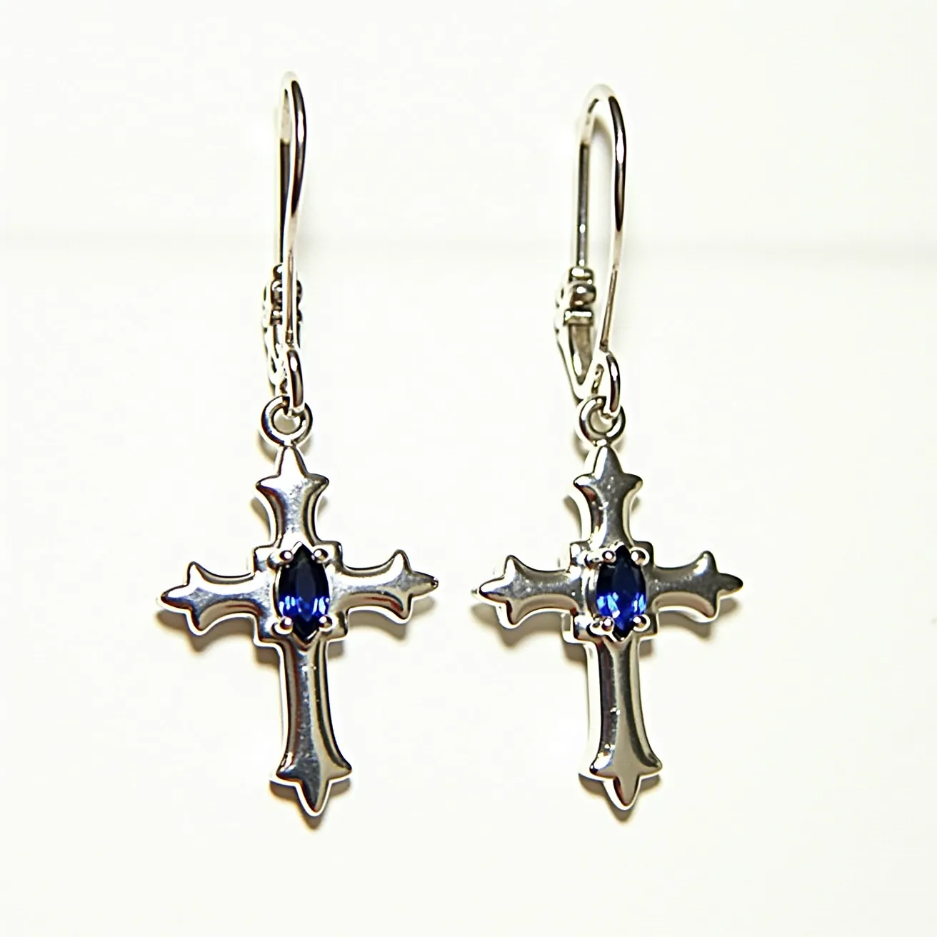 These cross earrings for men feature a shiny metal design with a prominent cross shape. At the center of each cross is an oval-cut blue gem, likely a sapphire, set in a four-prong setting that adds a touch of elegance and color contrast. The earrings are equipped with lever-back clasps, providing security and ease of wear. The overall design blends simplicity with a touch of sophistication, making them a versatile accessory.