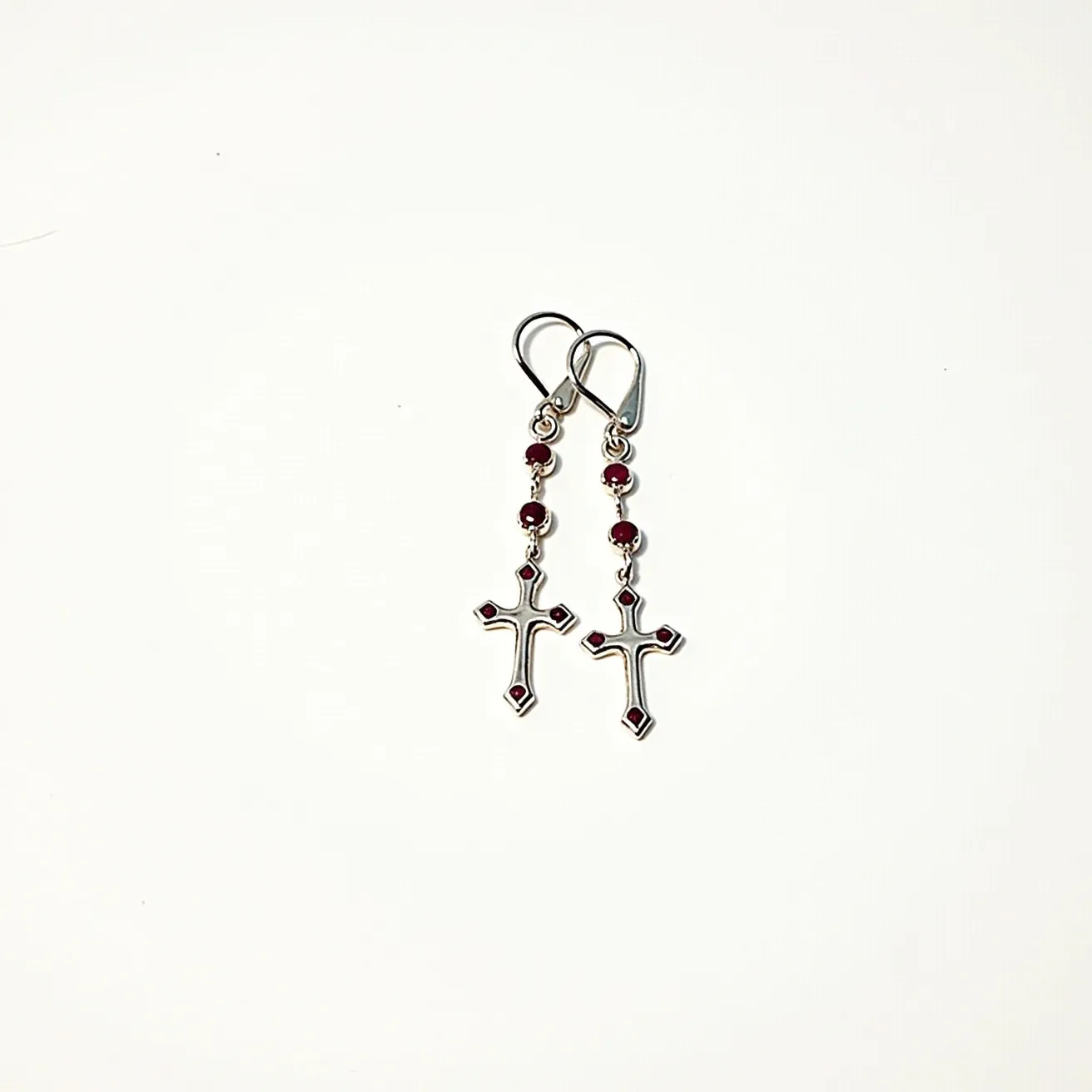 These cross earrings for men feature a silver-toned metal design with a distinct cross shape. Each cross is adorned with small, red gemstones positioned at the tips, showcasing a smooth round cut. The stones are bezel set, adding a subtle detail that complements the elegance of the earrings. The earrings are connected to small red beads via a slender chain, providing additional aesthetic appeal. They attach to the ear with a classic hook clasp, ensuring secure and comfortable wear.