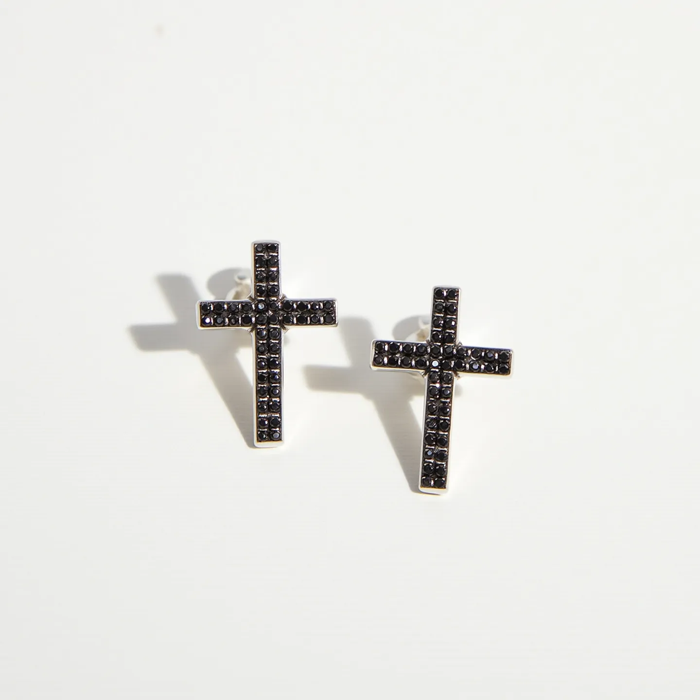 These cross earrings for men feature a striking design with numerous small, black stones set intricately along the entire cross, giving a sophisticated and edgy appearance. The stones are cut in a uniform shape, likely round, and are closely set in a pave style, allowing them to catch and reflect light effectively. The crosses are framed in what appears to be a silver metal, providing a polished contrast to the dark stones. The earrings are designed to be worn with a post-back attachment, ensuring a secure and comfortable fit for everyday wear.