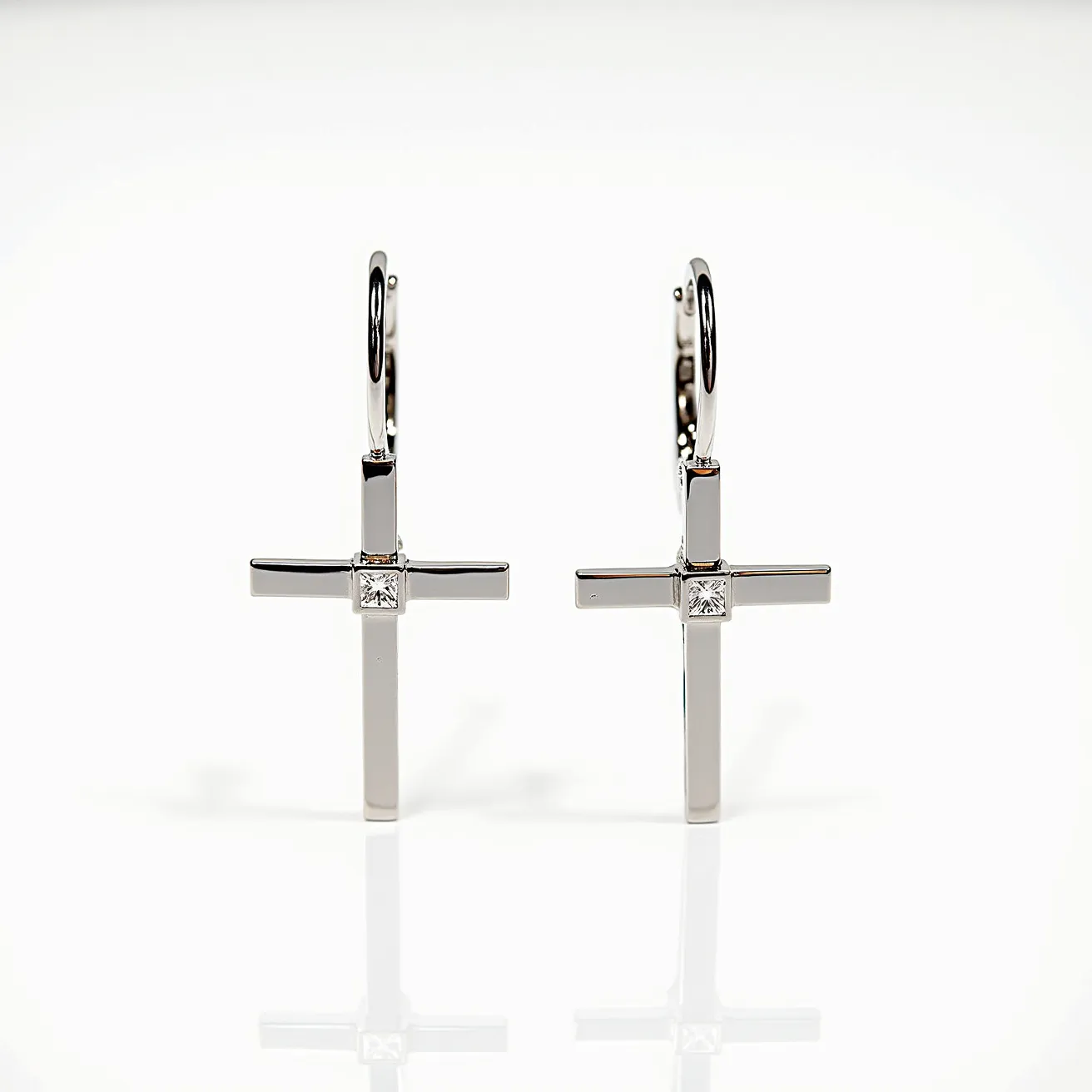 These cross earrings for men feature a sleek, metallic finish, likely crafted from a polished silver or white gold material. Each earring showcases a single square-cut gem set centrally in the cross, adding a touch of elegance and sparkle to the design. The stones appear to be mounted in a bezel or prong setting, providing security and prominence to the gems. The earrings are designed with a hoop clasp, allowing for a secure and comfortable attachment to the ear.