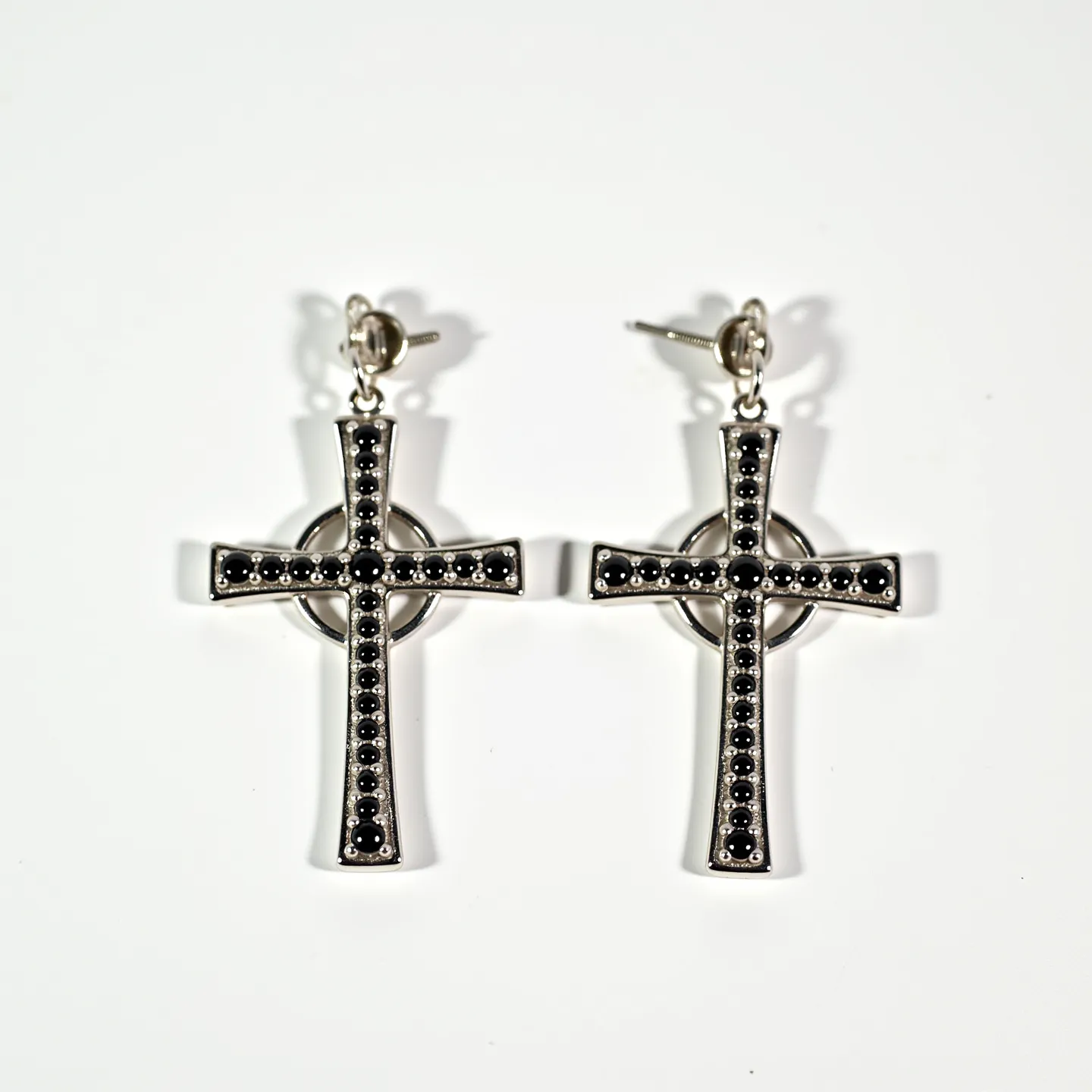 These cross earrings for men feature a sleek and metallic design, likely crafted from a silver-toned metal. Each earring displays a cross shape adorned with small, black round gems arranged in a linear pattern along the center of the cross. The stones appear to be faceted, providing a subtle sparkle, and are set into the metal using a prong setting. The earrings are attached via a stud-style clasp, comprising a post with a butterfly back, ensuring secure and comfortable wear.