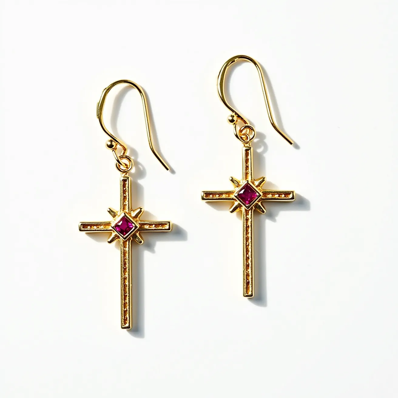 These cross earrings for women are elegantly crafted from a gold-toned material, giving them a polished and luxurious appearance. Each earring features a central gemstone that appears to be a red, square-cut gem, set within a starburst design at the intersection of the cross. The earrings are completed with a simple hook attachment, allowing for easy and secure wearing. Their intricate design and the vibrant color of the gemstone make them a striking accessory.