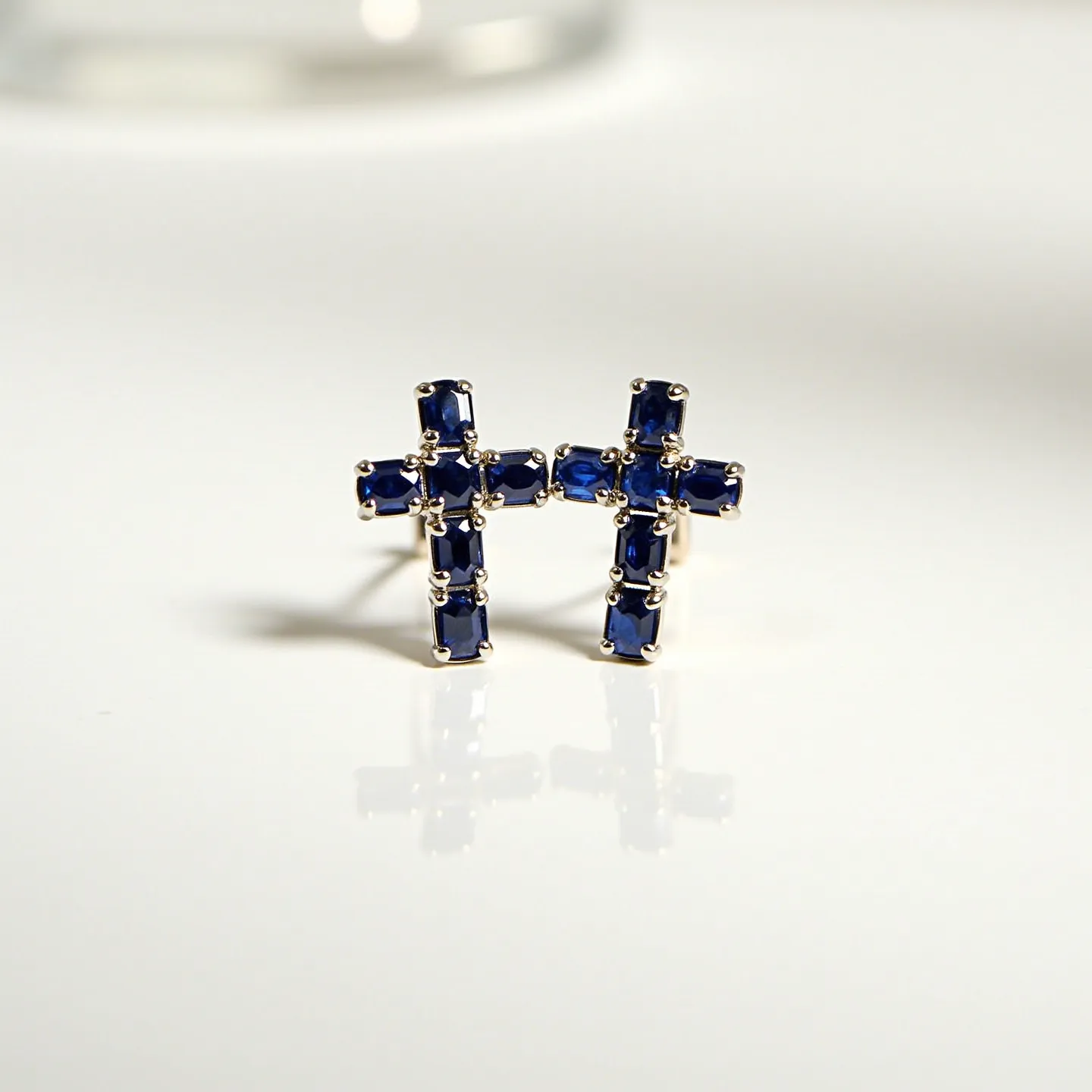 These cross earrings for women feature a design incorporating deep blue gemstones, likely sapphires, which are cut in a rectangular shape. Each stone is carefully set in a classic four-prong setting, ensuring security and stability. The gemstones form the recognizable silhouette of a cross, with three stones on each axis. The setting appears to be crafted from a bright metal, possibly white gold or platinum, providing a striking contrast to the dark blue stones. The earrings are secured with a post and butterfly clutch back, making them suitable for pierced ears and providing a comfortable fit.