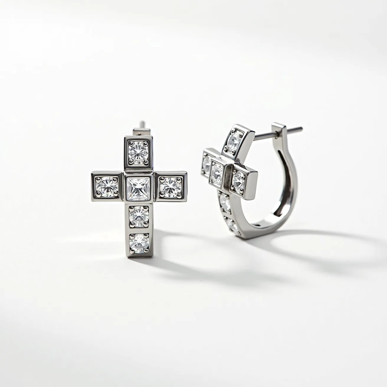 These cross earrings feature a sleek metallic design with a polished finish, likely made from silver or white gold. Each earring showcases a cross shape embellished with multiple clear, sparkling gems. The gems are primarily round-cut, but the center stone appears to be princess-cut, all set in a secure bezel setting that enhances their brilliance. The earrings have a hoop design with a post and clasp mechanism for easy and secure attachment. The overall design is elegant and sophisticated, adding a touch of sparkle.