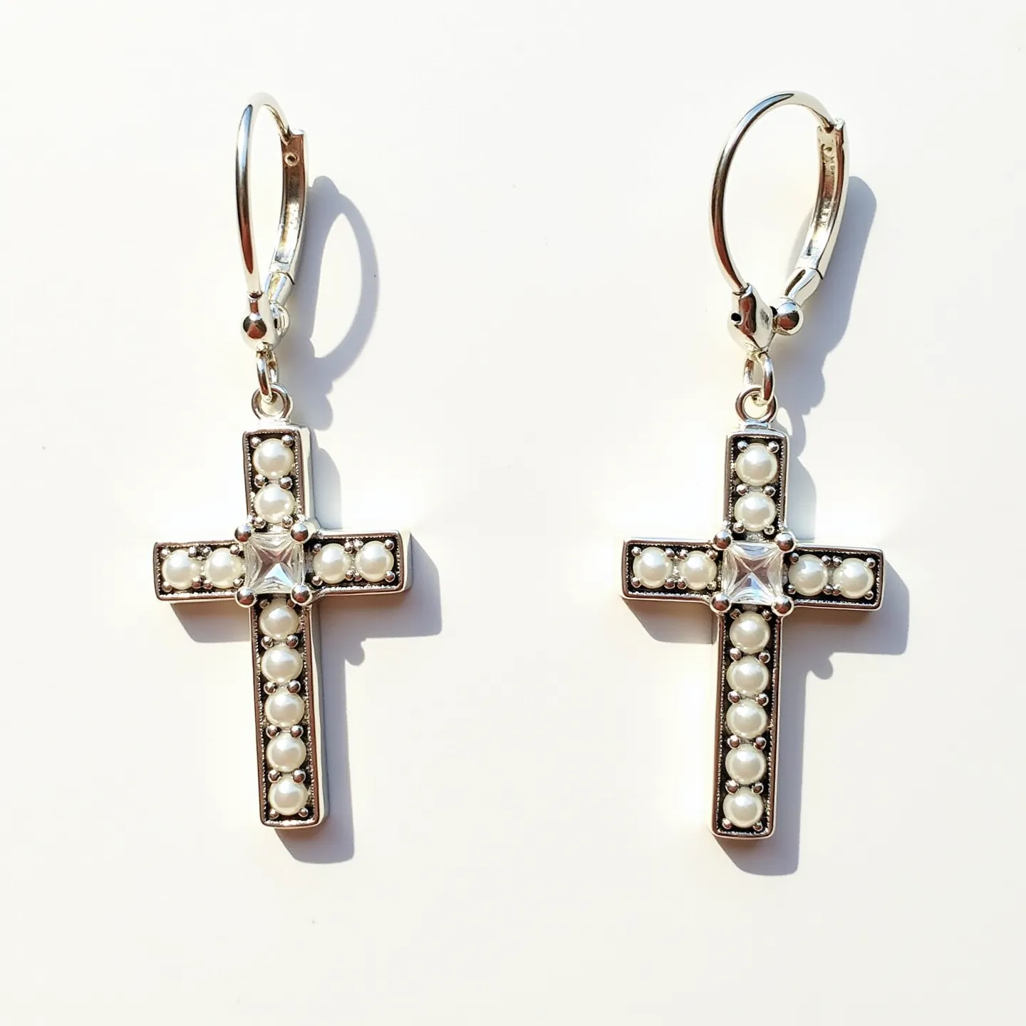 These cross earrings feature a well-crafted design made of a metallic material, possibly silver in color. Each earring is adorned with small, round pearls set along the length of the cross, adding a classic touch. At the center of the cross, there is a larger, square-cut gem, likely a clear crystal, held in place by four prongs. The earrings are attached to lever-back clasps, offering a secure and comfortable fit for the wearer. The overall design blends elegance with simplicity, making them a versatile accessory.