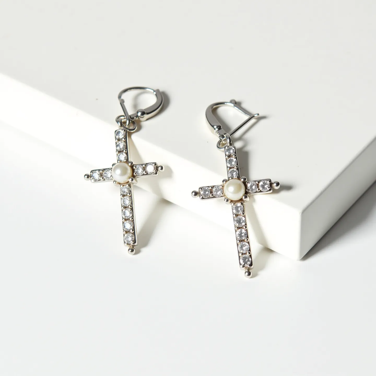 These cross earrings for women feature an elegant, metallic design, likely made from a polished silver or white gold material. They are adorned with small, round-cut clear stones, possibly diamonds or cubic zirconia, set in a pave style along the arms of the cross. At the center of each earring, a single round pearl is prominently positioned, adding a touch of sophistication. The earrings are attached with lever-back clasps, providing a secure and comfortable fit for the wearer.