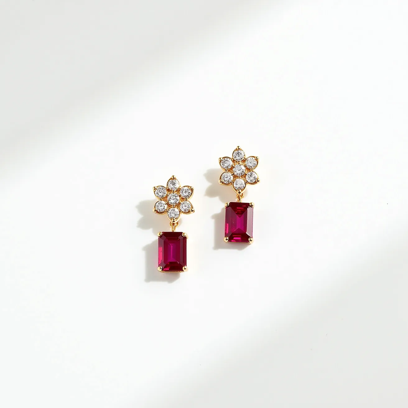 These dangle earrings are crafted with a delicate combination of materials, featuring a floral motif set with small, round diamonds that glimmer brightly. The petals of the flower are meticulously arranged around a central stone, all nestled within a finely detailed gold setting that adds to its elegance. Dangling below the floral design are rectangular step-cut stones, likely rubies, held securely in gold prong settings. The attachment is designed to gracefully hang, enhancing the movement and sparkle of both the diamonds and the richly colored stones beneath.