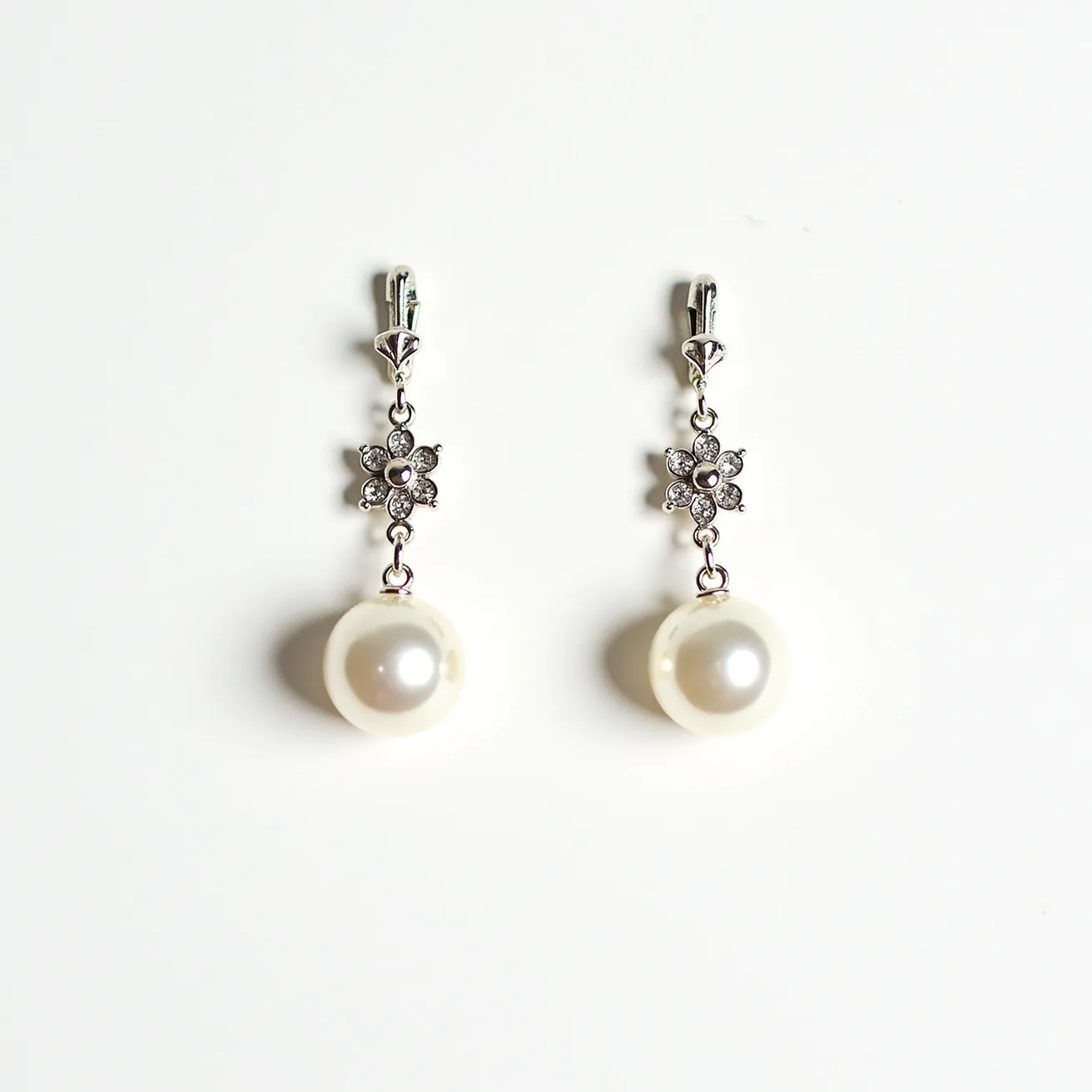 These dangle earrings feature a sophisticated design with a blend of materials. The earrings showcase a central luminescent pearl, smoothly polished, and round in shape. Above the pearl is a flower-like arrangement of small, round-cut sparkling stones, possibly diamonds or cubic zirconia, each set securely in prongs. The flower motif adds an elegant touch and provides a graceful transition between the pearl and the upper section of the earring. The earrings are attached via a sleek metallic clasp, likely silver or white gold, which ensures secure wearability. The harmonious combination of pearls and sparkling stones creates a timeless aesthetic suitable for various occasions.