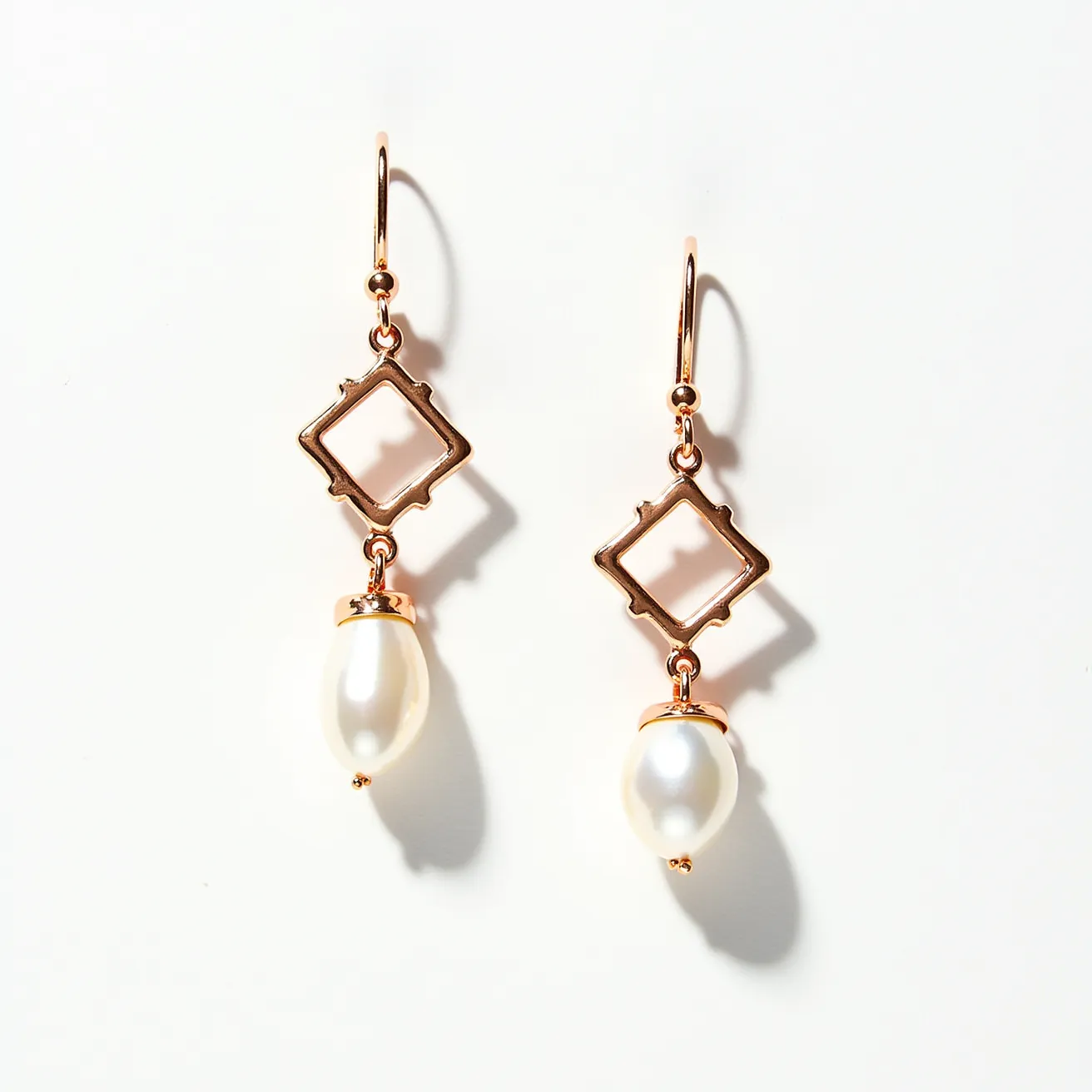 These dangle earrings feature a delicate combination of gold-toned metal and white pearls. The design includes an open square shape crafted from metal, providing an elegant, geometric focal point. Beneath each square, a white teardrop-shaped pearl is suspended, adding a classic charm to the modern geometric design. The pearls are likely set in a simple peg setting, allowing them to sway freely and catch the light. The attachment features a classic fishhook clasp, providing both security and ease of wear.