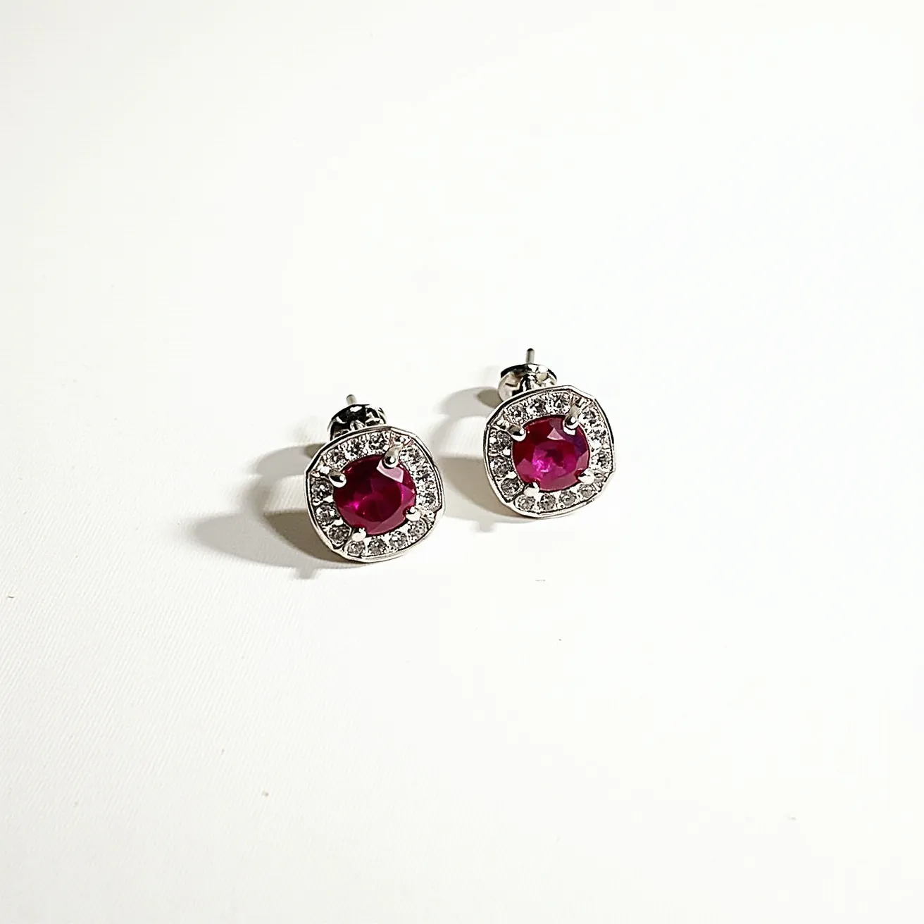 These diamond earrings feature a central round-cut red gemstone set in a prong setting, likely a ruby given its vibrant color. Surrounding the central gem is a halo of small round diamonds, each meticulously placed in a pavé setting that adds extra brilliance. The earrings have a metal base, possibly white gold or platinum, which enhances the sparkle of the stones. A classic butterfly back serves as the attachment, ensuring a secure and comfortable fit on the ear. The elegant combination of red and clear gemstones creates a stunning contrast, making these earrings a sophisticated accessory for any occasion.