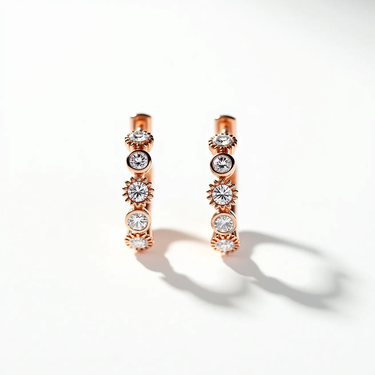 These diamond hoop earrings are crafted from a lustrous metal, likely rose gold, showcasing a series of round brilliant-cut diamonds. Each diamond is elegantly set in a bezel-like setting, framed by intricate, starburst details that add a touch of elegance and enhance the overall brilliance of the gemstones. The continuous design of the hoops suggests they secure with a hidden hinge or clasp mechanism, ensuring a seamless appearance when worn. The use of high-quality materials and careful craftsmanship is evident in the refined and sophisticated look of these earrings.