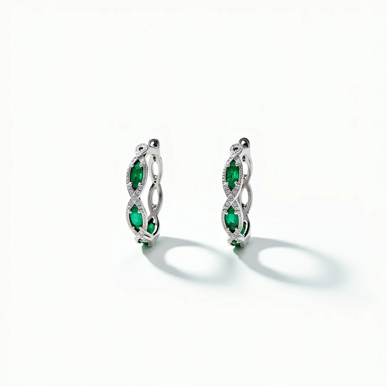 These diamond hoop earrings feature a stunning design crafted from what appears to be white gold or platinum. Each earring is adorned with vibrant green oval-cut stones, likely emeralds, set between intricate twirls of small round-cut diamonds that enhance their sparkle. The diamonds are arranged in an elegant crossover pattern, adding to the sophistication of the hoops. The earrings are equipped with secure hinge-back clasps, ensuring both elegance and practicality for the wearer.