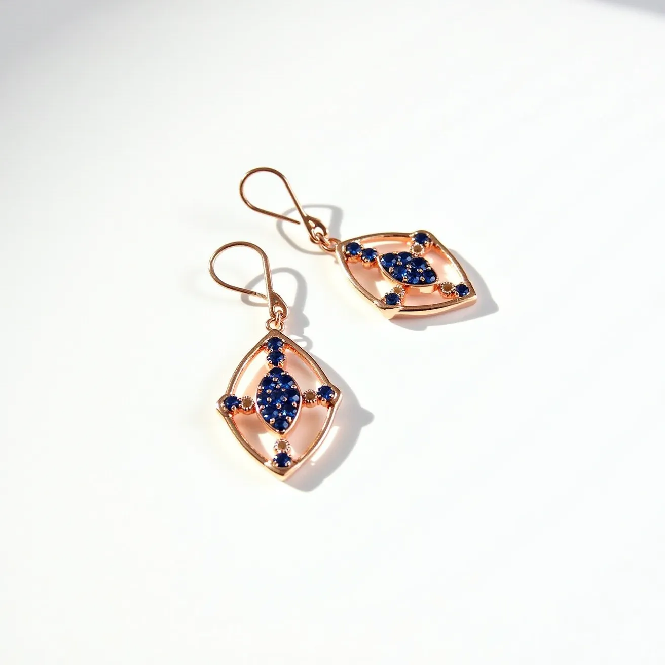 These drop earrings feature an elegant, elongated diamond shape made from a rose gold metal that beautifully complements the deep blue gems adorning each piece. The gems, likely sapphires, are arranged in a captivating marquise cut at the center, with smaller round stones enhancing the points of the diamond-shaped frame. The intricate setting allows the stones to catch the light, adding a sparkling contrast to the warm tone of the gold. The earrings are suspended from simple hook clasps, ensuring a secure and comfortable fit while maintaining a sleek and sophisticated aesthetic.