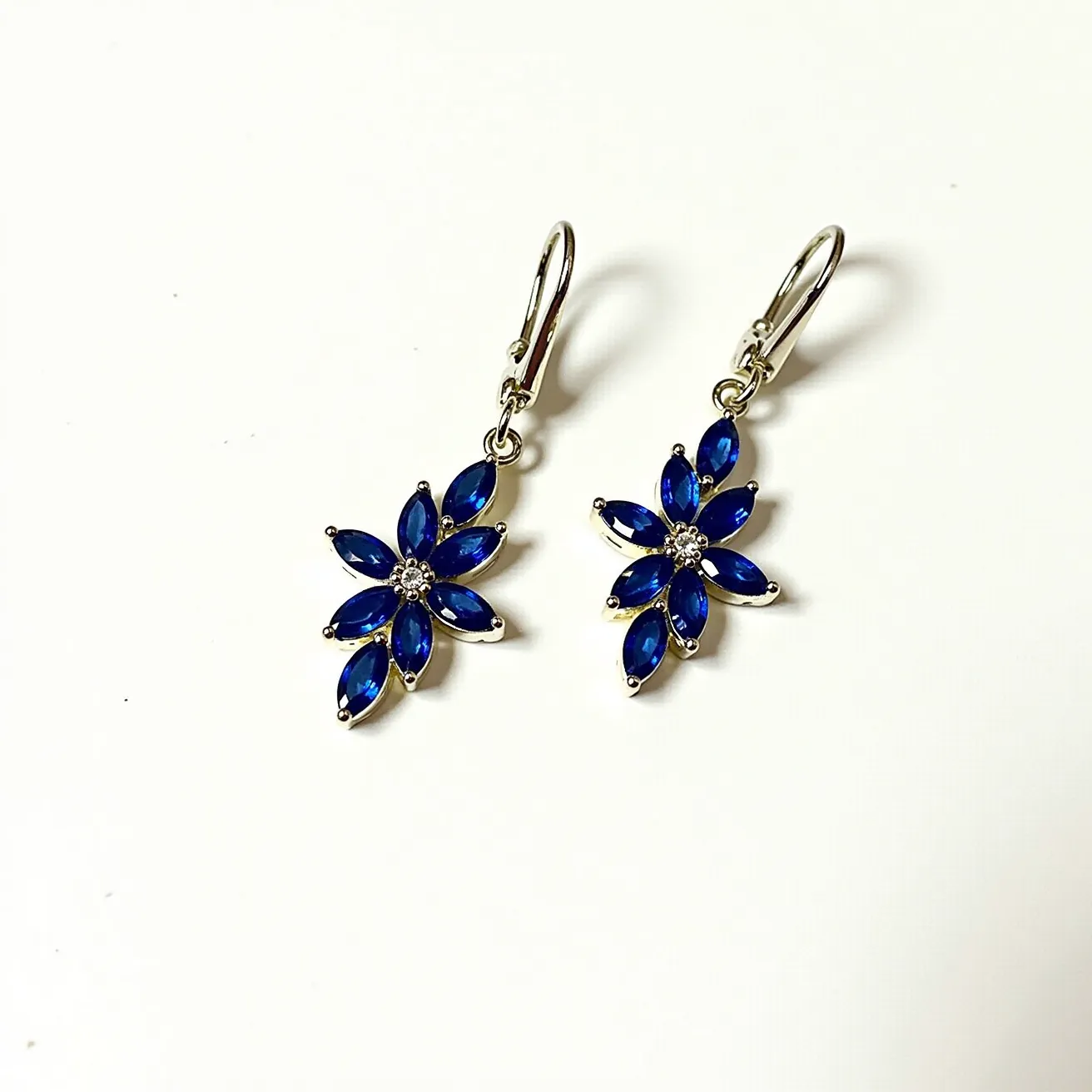 These drop earrings showcase a floral design featuring marquise-cut blue stones set in a brilliant flower pattern. Each earring is accented by small, clear stones at the center, adding a touch of sparkle. The metal used appears to be a polished silver or white gold, enhancing the vibrant blue of the stones. These earrings are attached with lever-back clasps, ensuring secure and comfortable wear. The overall craftsmanship highlights elegance and a classic appeal, perfect for both formal and casual occasions.