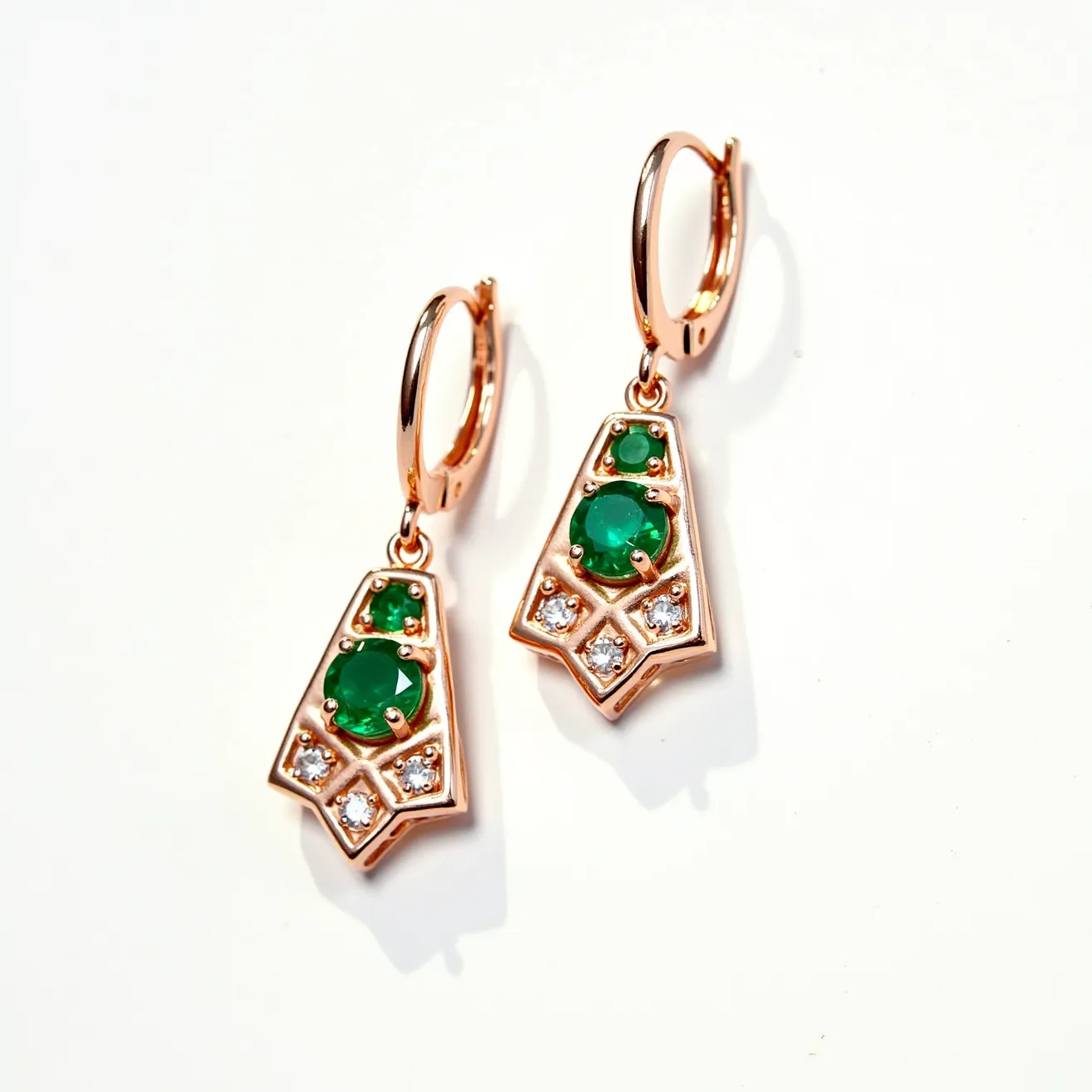 These emerald earrings feature an exquisite combination of vibrant green emeralds, intricately set in a rose gold metal framework. The primary gemstones are round-cut emeralds, with the larger central emerald surrounded by smaller ones, enhancing their rich color. The design is complemented by white, round-cut diamonds set in a geometric pattern, adding brilliance and contrast to the overall appearance. The earrings have a secure hinged hoop clasp that ensures ease of wear while maintaining an elegant drop design.