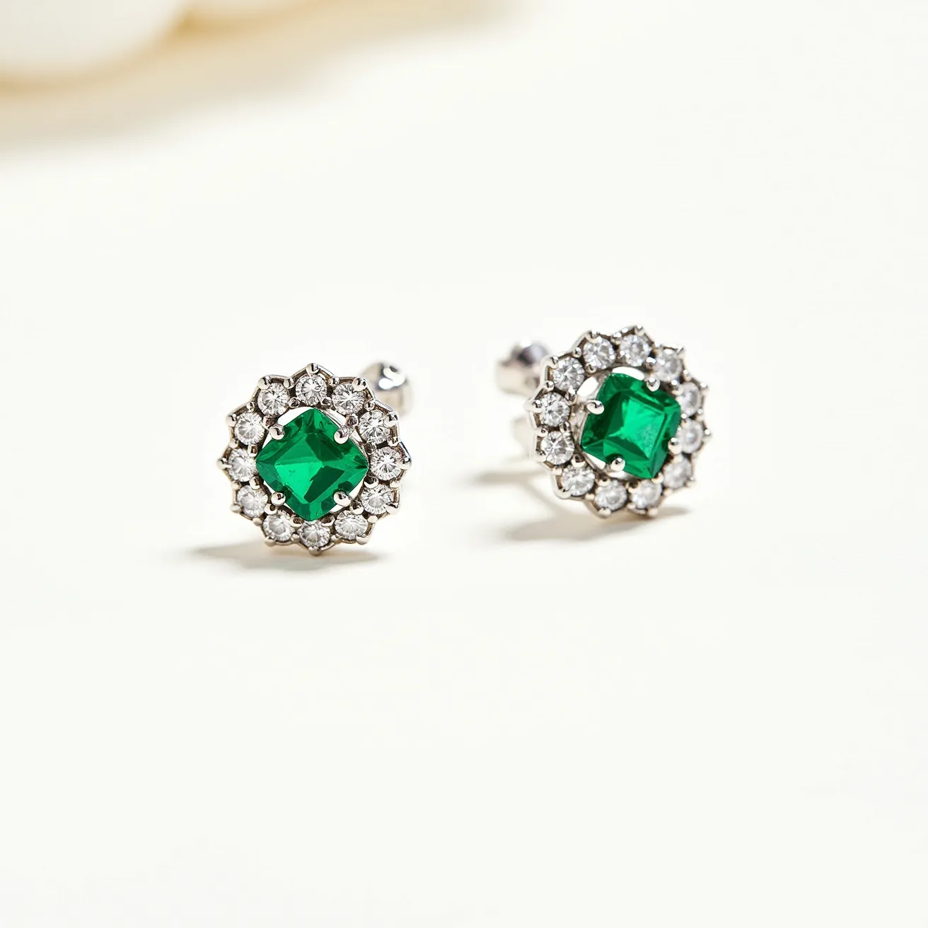 These emerald earrings feature a central, square-cut emerald gemstone, surrounded by a halo of smaller round-cut white diamonds, enhancing the vibrant green hue of the primary stone. The gemstones are set in a prong setting, allowing each gem to be showcased prominently. The earrings have a sleek metal base, likely white gold or platinum, contributing to their elegant and luxurious appearance. A secure post and butterfly clasp attachment ensures that the earrings stay comfortably in place when worn, blending style with practicality.