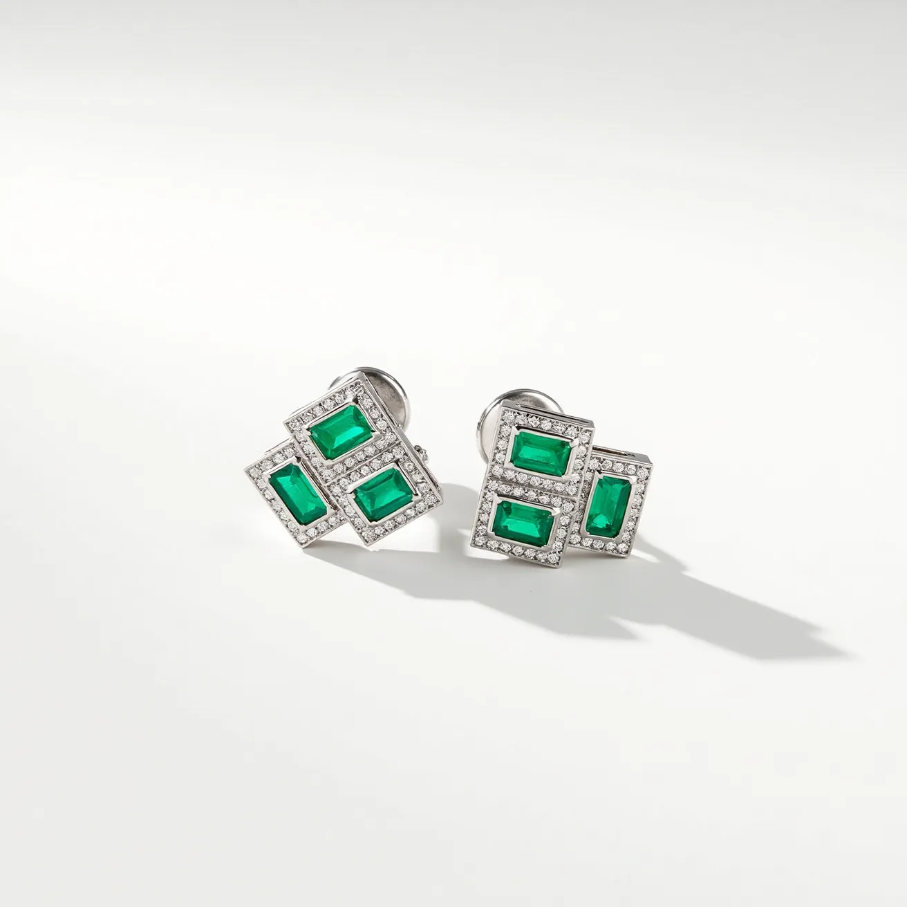 These emerald earrings feature a striking design with each earring showcasing four vibrant green emeralds in rectangular cuts. The emeralds are set within a geometric arrangement, bordered by a halo of small, sparkling diamonds, adding elegance and contrast to the overall design. The materials suggest a high-quality metal, likely white gold or platinum, complementing the luminous green stones and enhancing the brilliance of the diamonds. These earrings have secure post attachments, ensuring they are both stylish and functional. The craftsmanship highlights the beauty of the gemstones while maintaining a sophisticated and luxurious appearance.