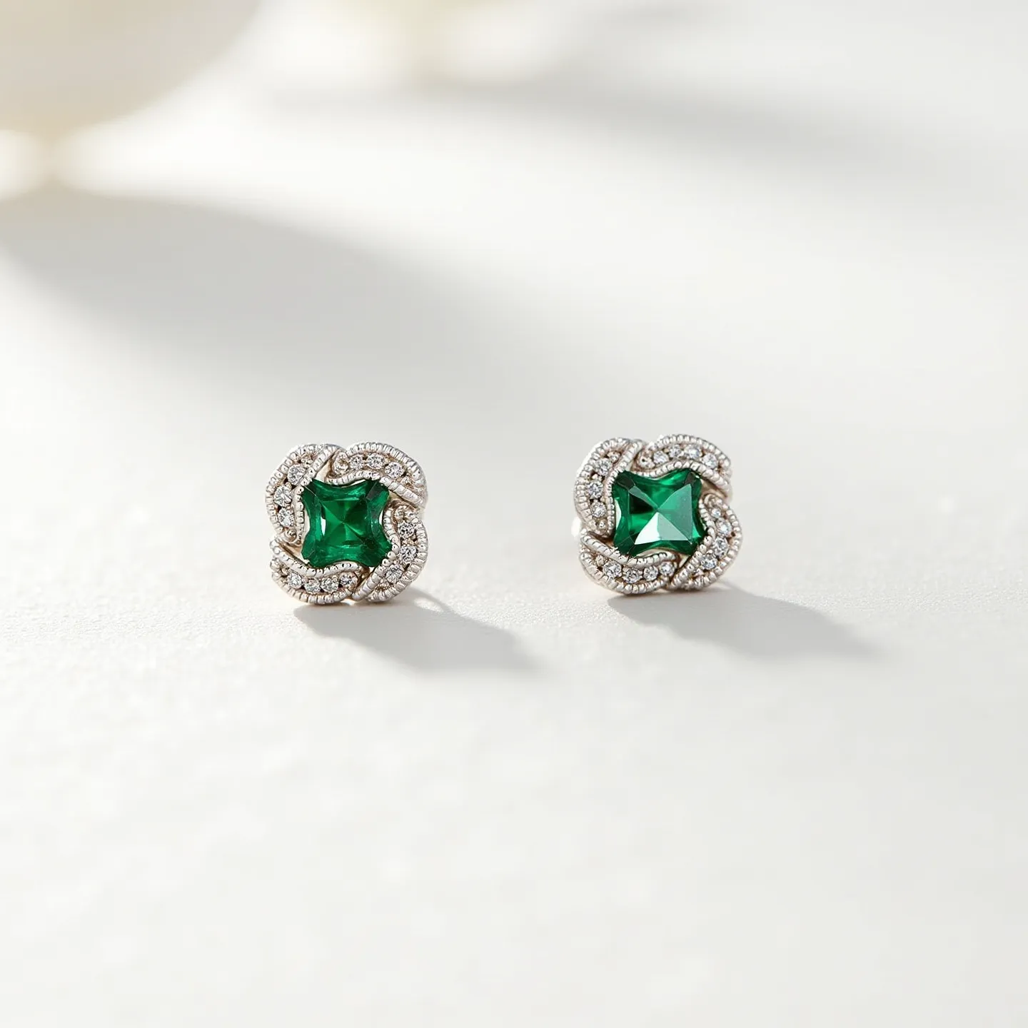 These emerald earrings display a captivating design featuring a central square-cut emerald, known for its vivid green hue. The emerald is elegantly framed by a halo of smaller clear gemstones, likely diamonds, which enhance its brilliance and sophistication. The setting appears to be crafted from a high-polish metal, such as white gold or platinum, providing a striking contrast to the vibrant green of the emerald. The earrings utilize a classic post and clutch back attachment, ensuring both a secure fit and comfortable wear. The intricate detailing in the setting adds a touch of timeless elegance, perfect for both formal and special occasions.