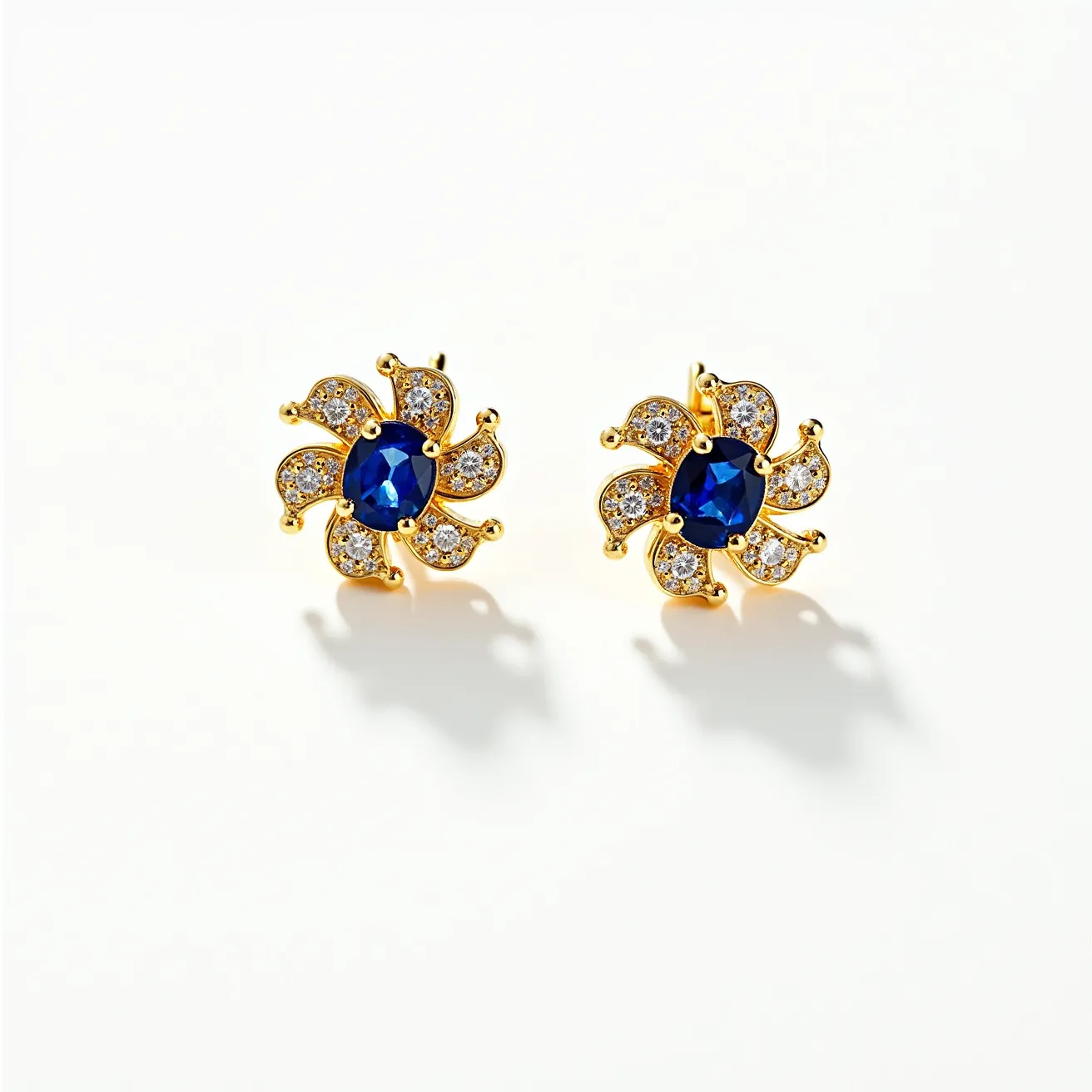 These floral earrings showcase a stunning design featuring a central blue gemstone, shaped in an oval cut and set securely in the center. Surrounding the central gem are gold-toned petal-like elements, each adorned with small sparkling stones that resemble diamonds, set in what appears to be a pavé style. The overall effect is reminiscent of a blooming flower, with the rich blue stone serving as its heart. The earrings likely feature a post and butterfly clasp attachment, providing a secure and elegant fit. The combination of the deep blue center stone, the shimmering clear stones, and the warm gold tone creates a luxurious and vibrant aesthetic.